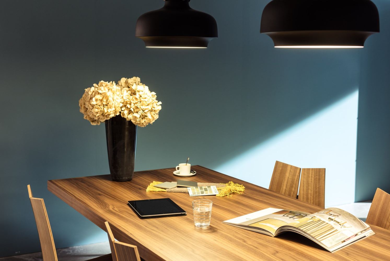 Holztisch unter zwei schwarzen Pendelleuchten vor blauem Wandfeld; darauf eine hohe Vase mit hellen Hortensien, Muster- und Farbfächer, Espressotasse mit Löffel, Tablet, Wasserglas und ein aufgeschlagenes Magazin, umgeben von vier Holzstühlen im Streiflicht.