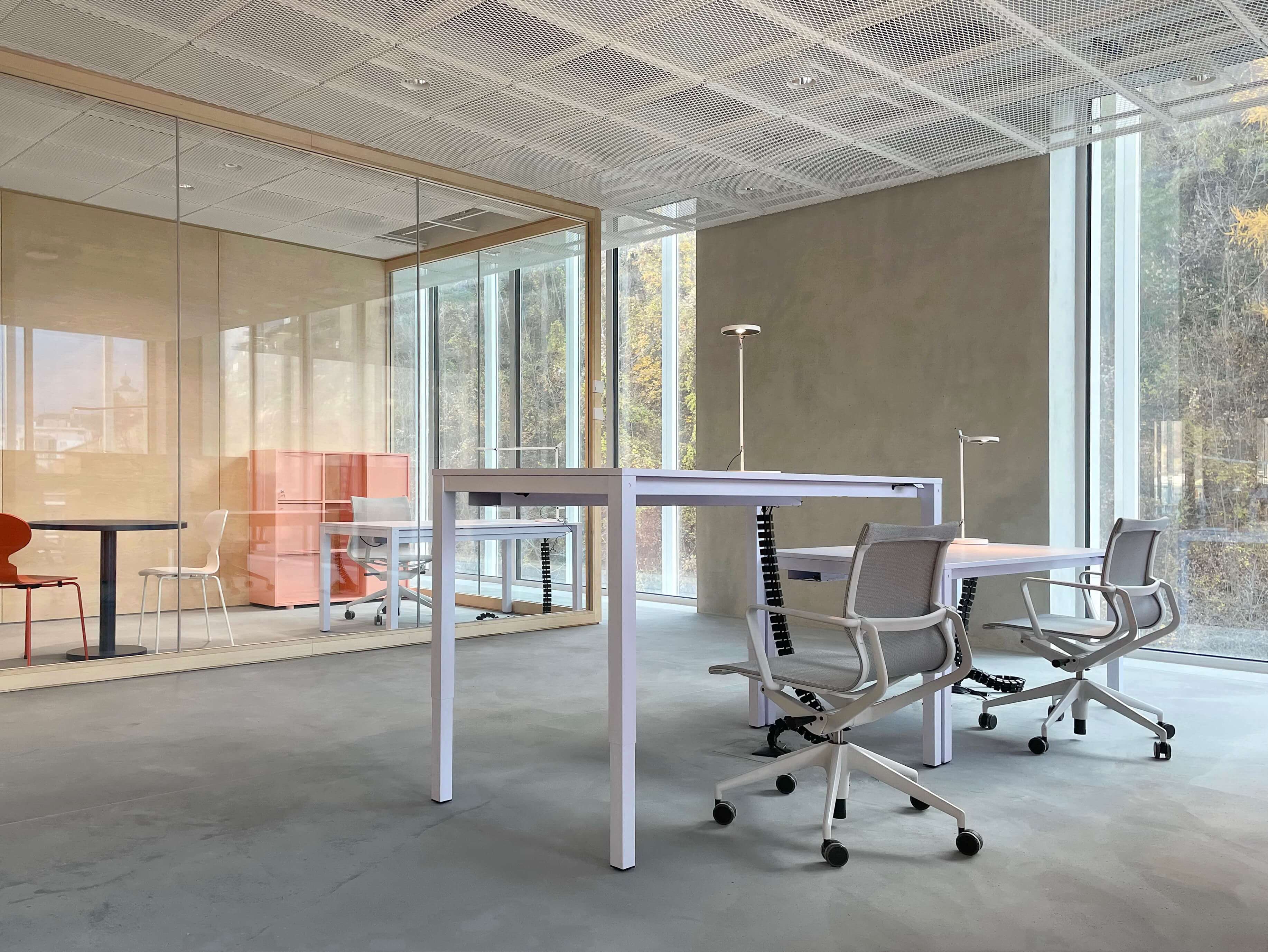 Arbeitszone mit höhenverstellbarem Tisch, Drehstühlen und Leuchten vor verglastem Besprechungsraum im Hochschulcampus in Brig.