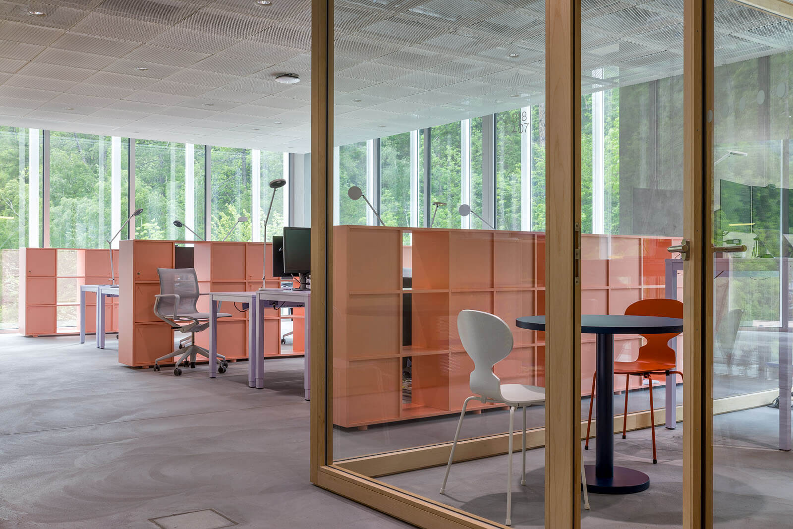 Glasbox mit rundem Besprechungstisch und Stühlen, dahinter offene Arbeitsplätze mit Leuchten im Hochschulcampus Brig.