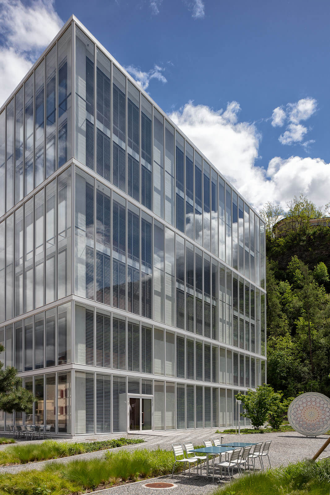 Gläserner Gebäuderiegel mit feinen Lamellen, davor Kieswege, Bepflanzung und ein Aussentisch mit Stühlen im Hochschulcampus in Brig.