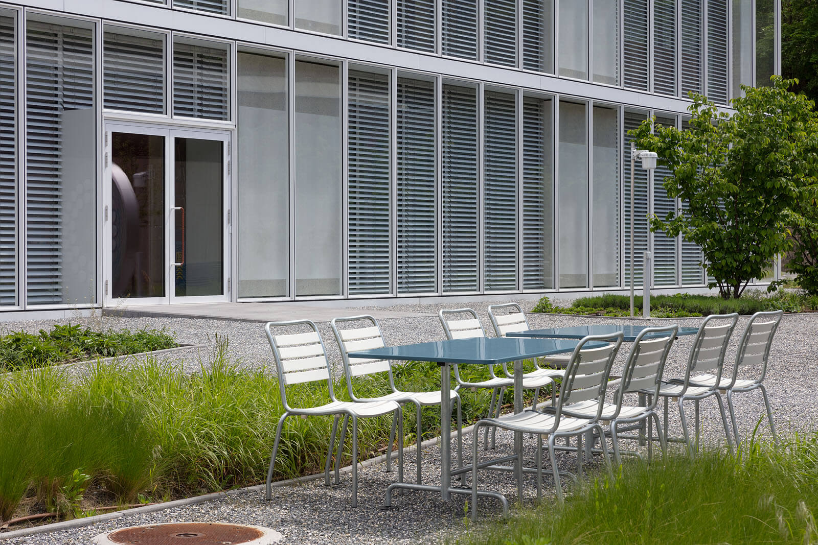 Aussenbereich mit langem blauen Tisch und weissen Metallstühlen vor der lamellenverkleideten Glasfassade im Hochschulcampus in Brig.
