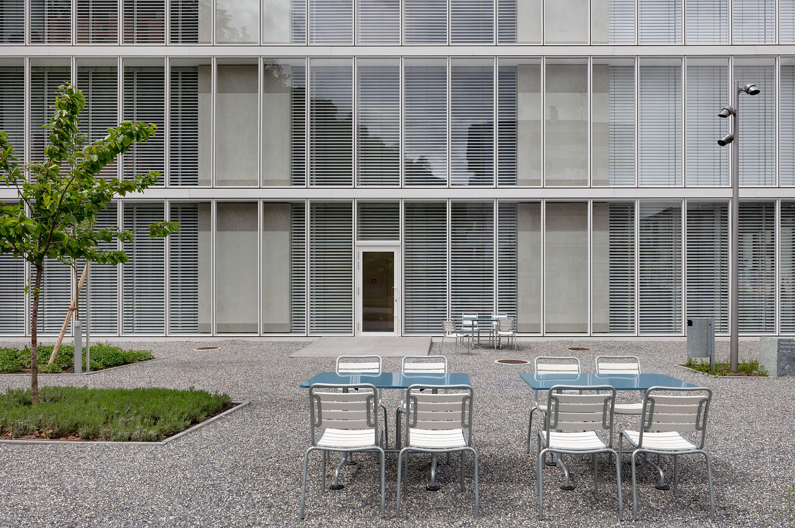 Innenhof mit zwei blauen Tischen und weissen Stühlen vor der rhythmischen Glas-Lamellenfassade im Hochschulcampus in Brig.