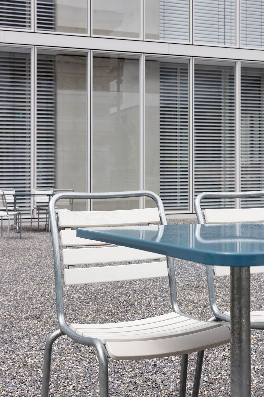 Nahaufnahme von blauem Aussentisch mit weissen Metallstühlen vor Glasfassade mit Lamellen im Hochschulcampus in Brig.
