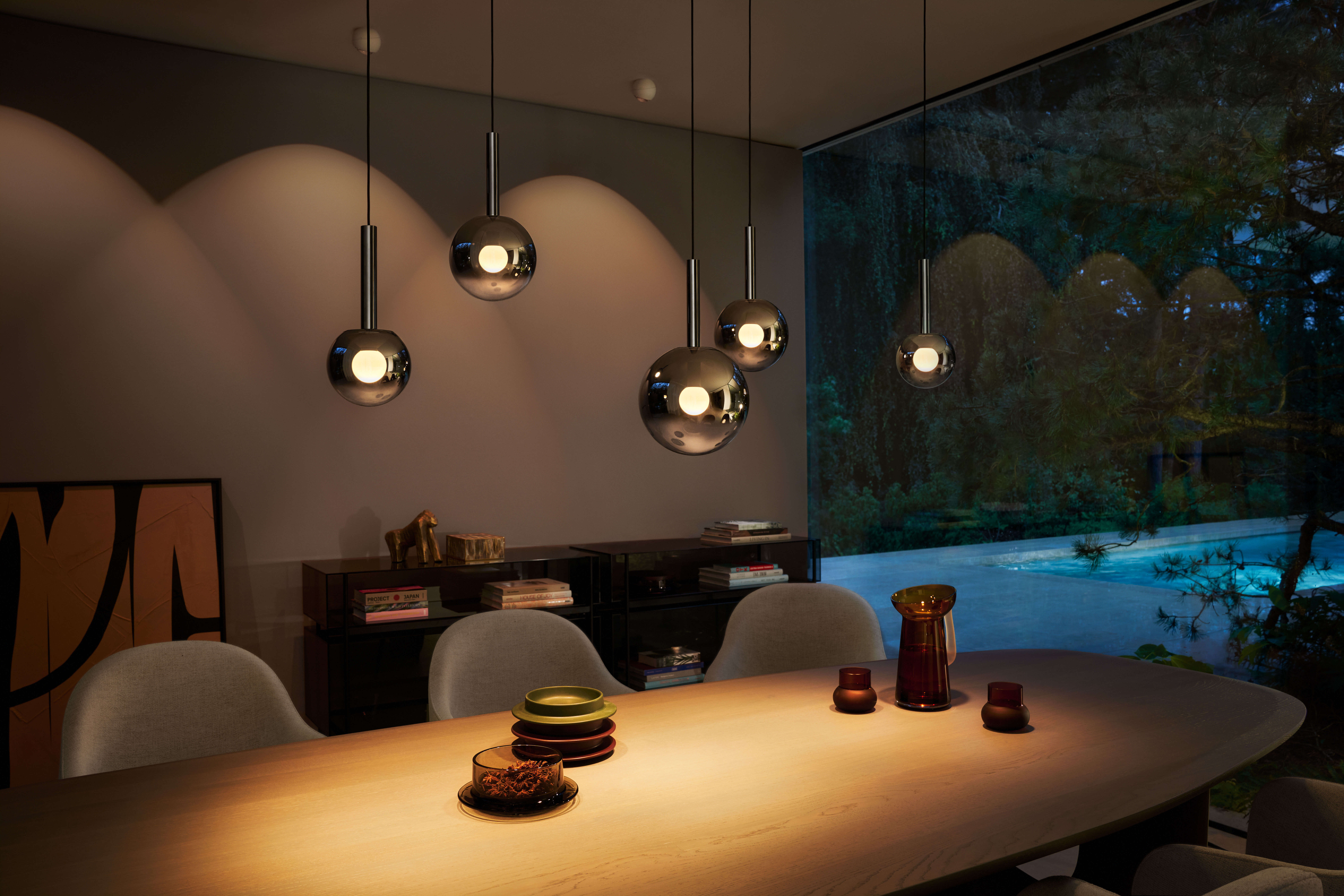 Wohn-/Essbereich: schwebende kugelfoermige Pendelleuchten über Holztisch, stimmungsvolles Abendlicht, Blick durch Panoramafenster in den Garten mit Pool.