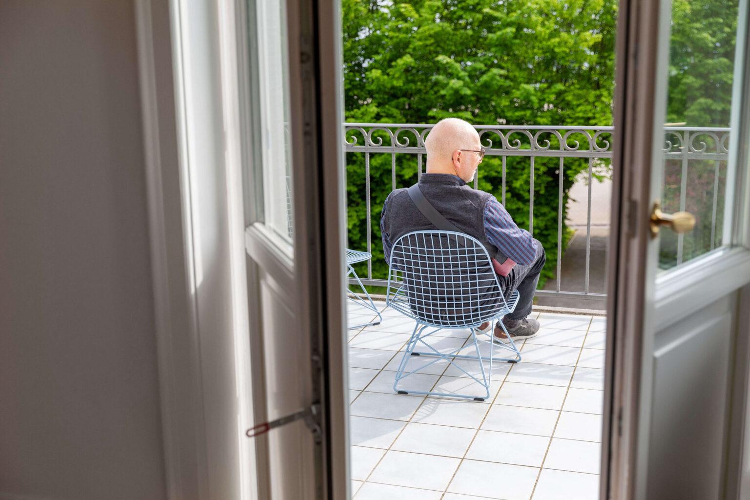 Mann sitzt auf Balkon in Drahtstuhl mit Blick ins Grüne.