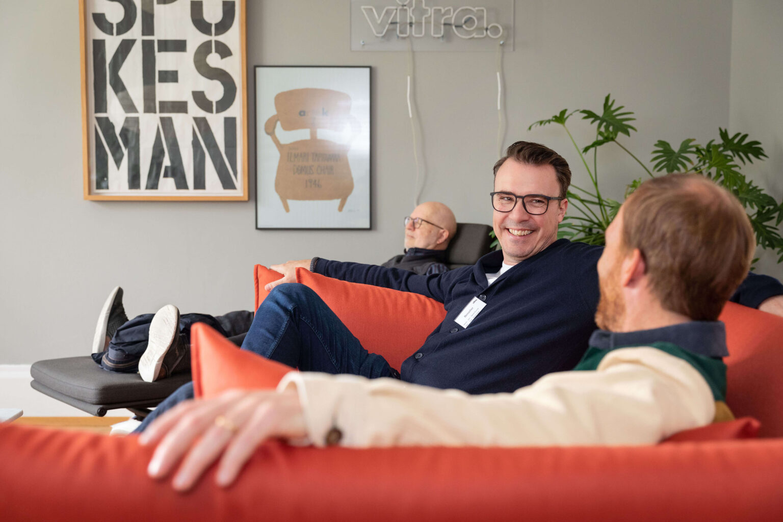 Drei Männer sitzen entspannt auf rotem Sofa im Büro.
