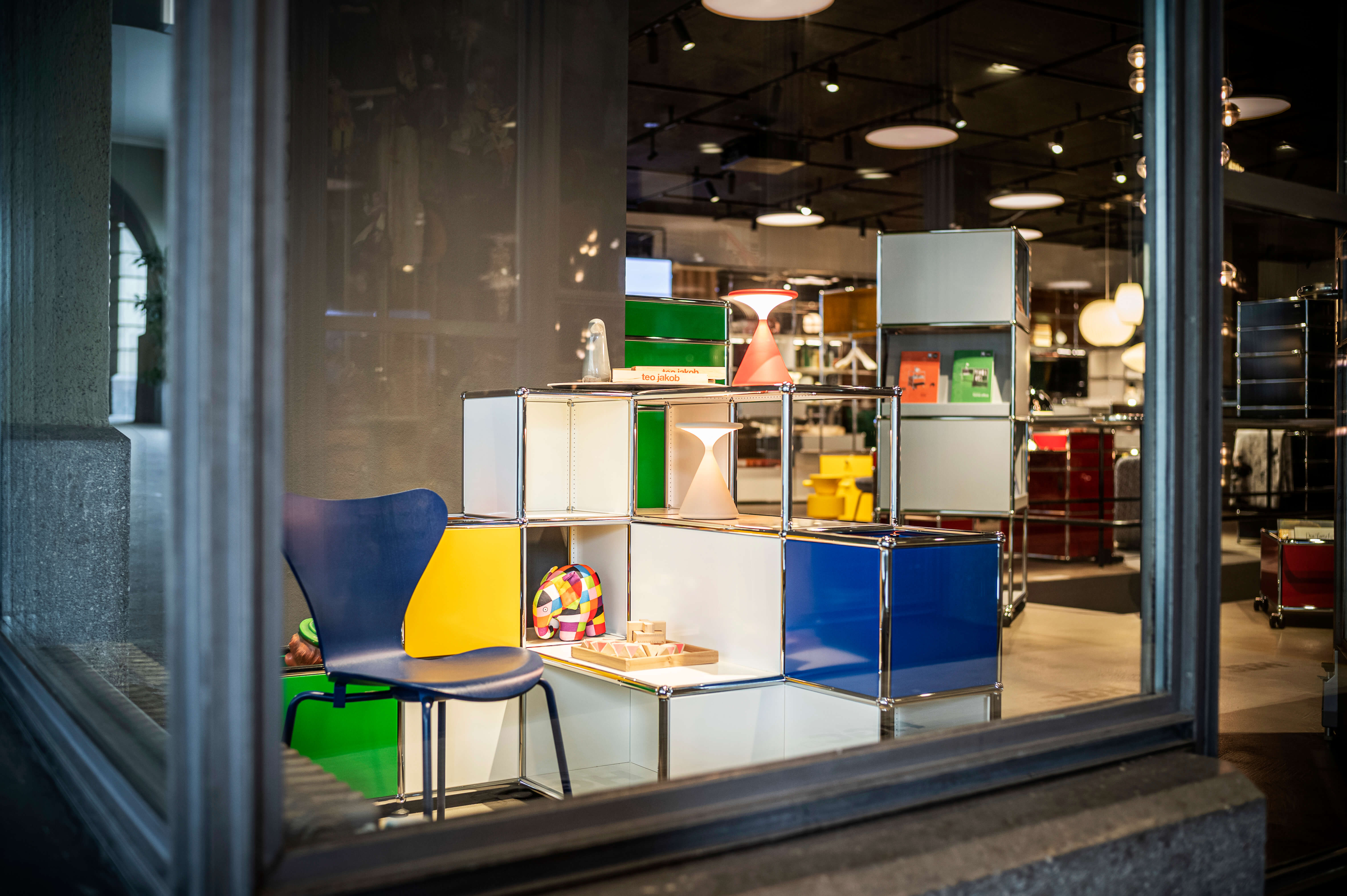Schaufensteransicht eines Showrooms mit modularen Regalelementen in Weiss, Blau, Grün und Gelb. Auf den Ablagen stehen kleine Leuchten und Dekoobjekte. Links ein blauer Stuhl. Im Hintergrund sind weitere Möbel und Pendelleuchten im Laden sichtbar.