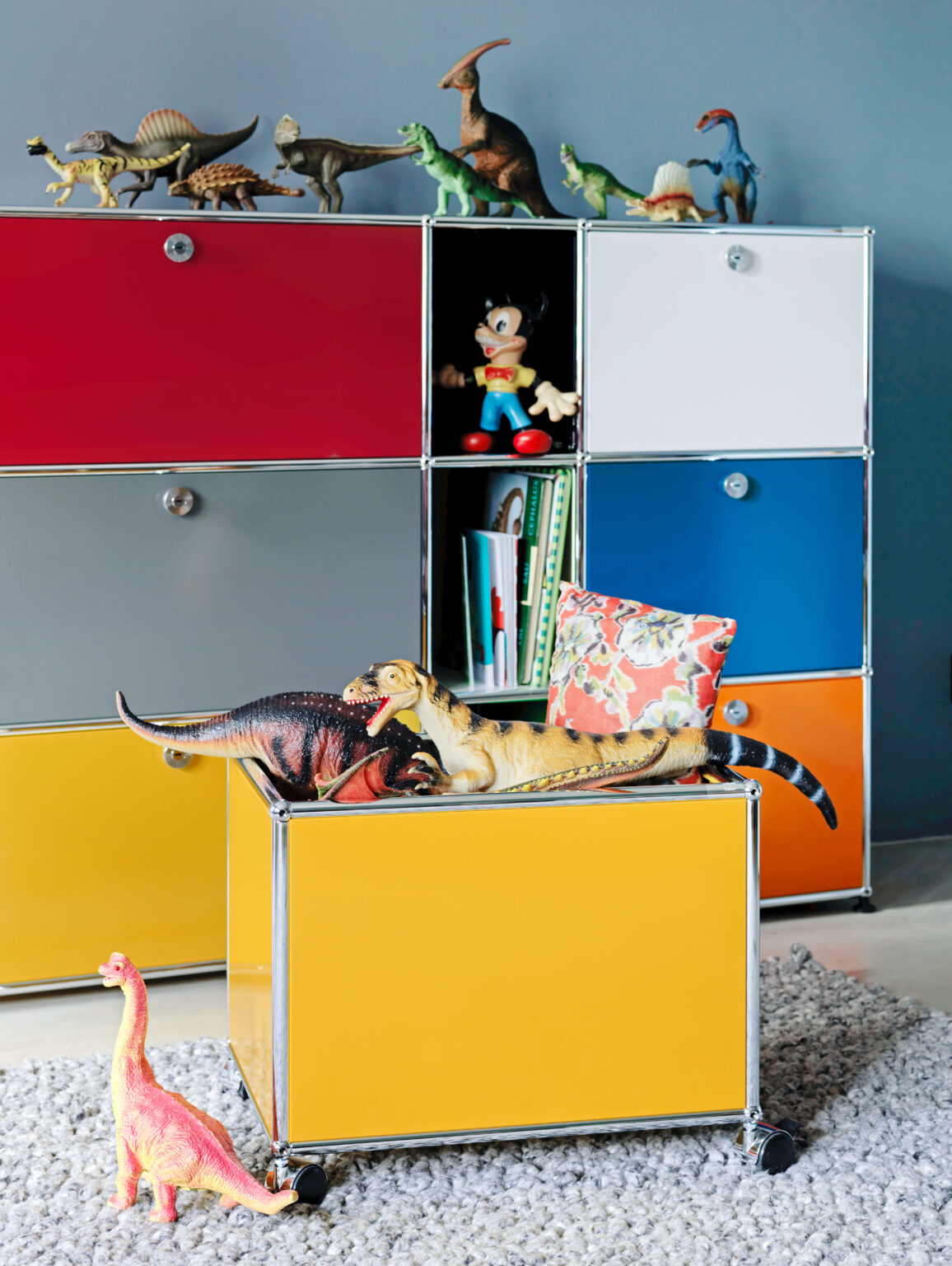 Buntes Aufbewahrungsmöbel mit farbigen Türen in Rot, Grau, Weiss, Blau, Orange und Gelb. In einem offenen Fach stehen Bücher und eine Spielzeugfigur. Auf dem Möbel und auf einem gelben Rollcontainer liegen mehrere Dinosaurier Figuren, davor steht ein kleiner rosa Dino auf einem Teppich.
