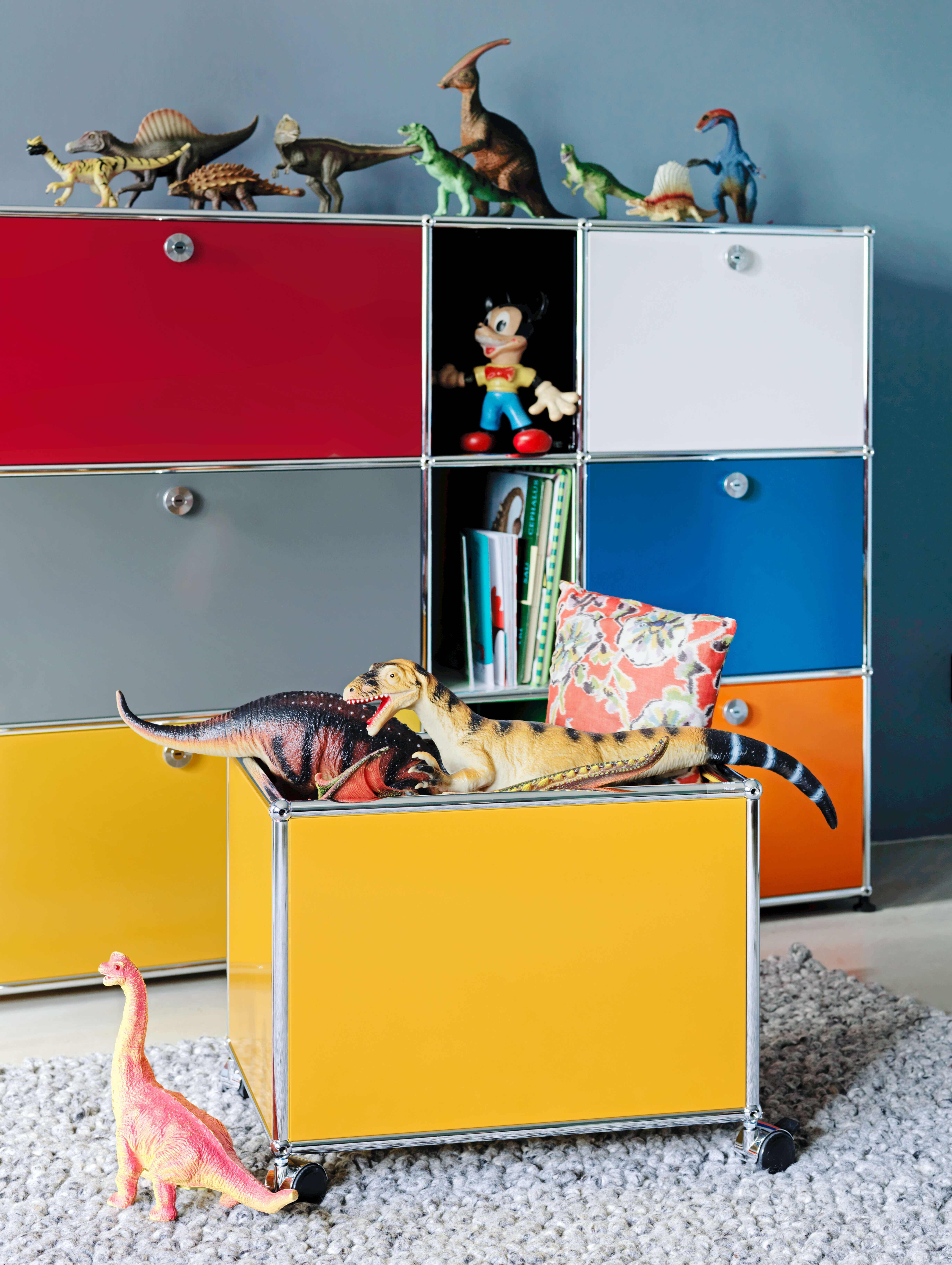Buntes Aufbewahrungsmöbel mit farbigen Türen in Rot, Grau, Weiss, Blau, Orange und Gelb. In einem offenen Fach stehen Bücher und eine Spielzeugfigur. Auf dem Möbel und auf einem gelben Rollcontainer liegen mehrere Dinosaurier Figuren, davor steht ein kleiner rosa Dino auf einem Teppich.