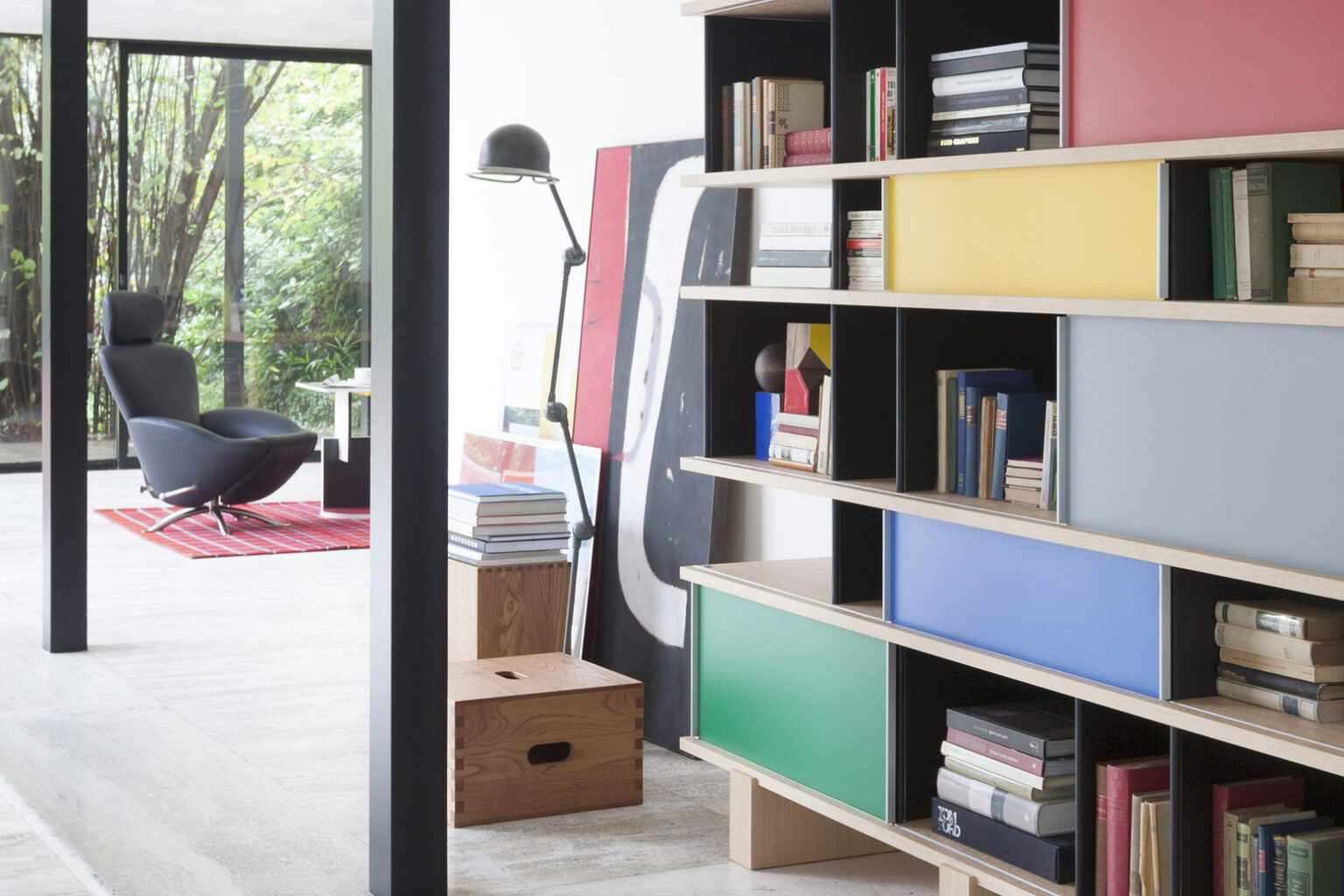 Modulares Regal mit offenen Fächern, Büchern und farbigen Schiebeelementen in Gelb, Blau und Grün. Im Hintergrund führt eine offene Durchsicht in einen hellen Raum mit schwarzem Lounge Sessel auf rotem Teppich, Stehleuchte und grossen Fensterfronten ins Grüne.
