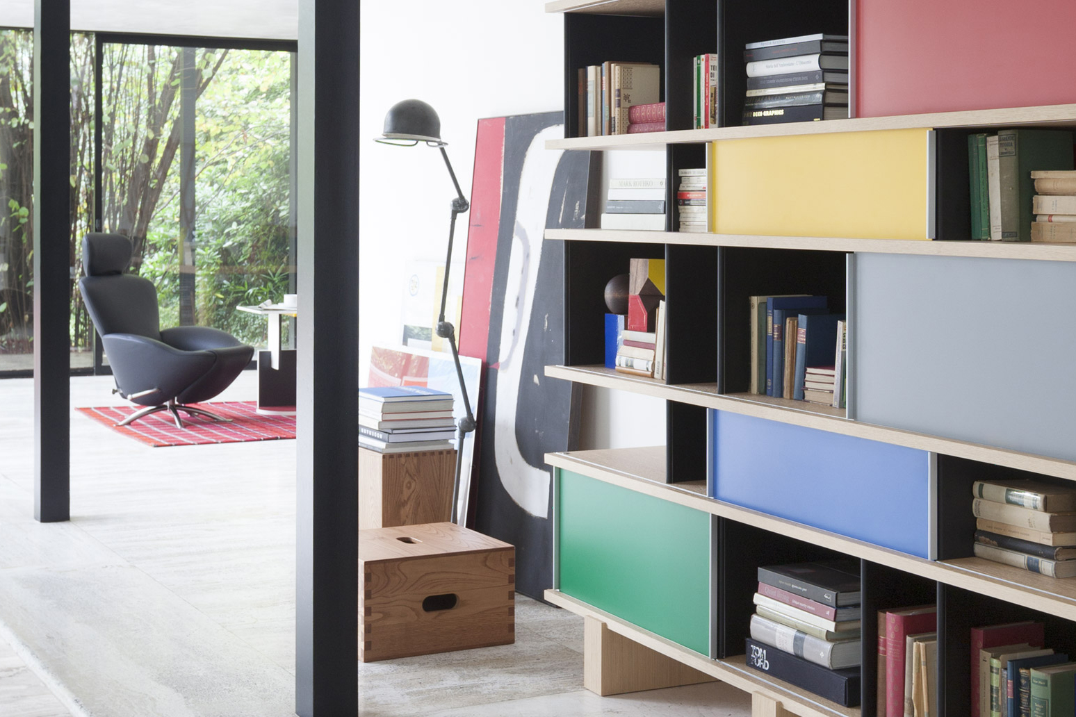 Modulares Regal mit offenen Fächern, Büchern und farbigen Schiebeelementen in Gelb, Blau und Grün. Im Hintergrund führt eine offene Durchsicht in einen hellen Raum mit schwarzem Lounge Sessel auf rotem Teppich, Stehleuchte und grossen Fensterfronten ins Grüne.