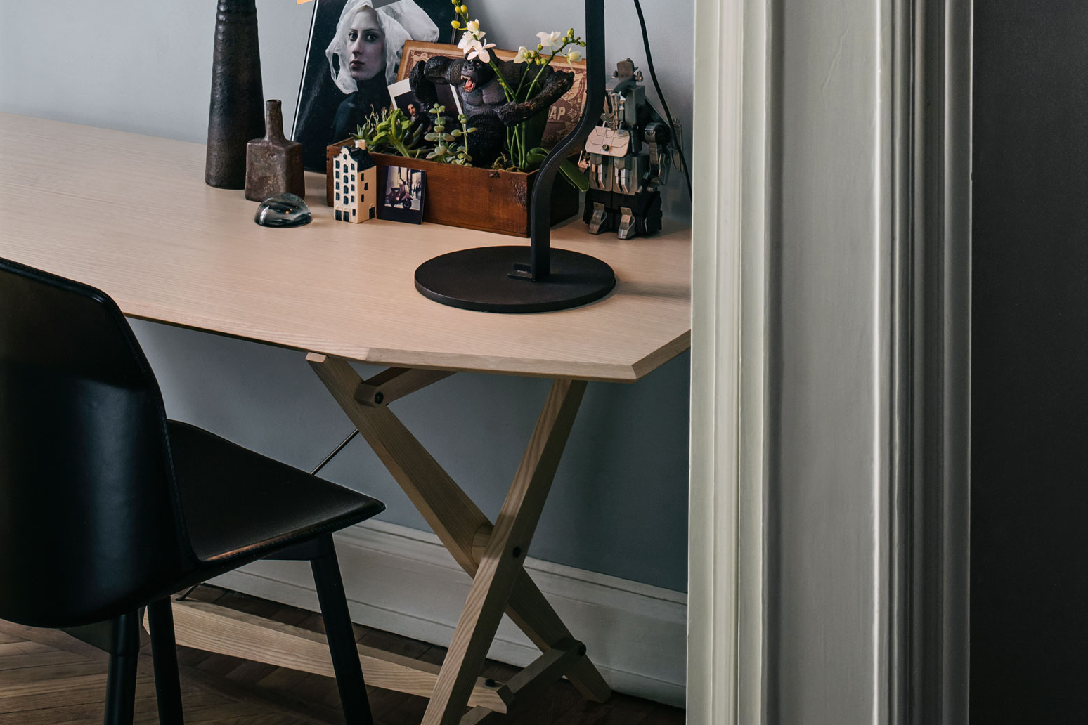 Schmaler Arbeitstisch aus hellem Holz mit X Gestell in einer Nische. Auf der Tischplatte stehen eine schwarze Tischleuchte, ein Tablett mit Pflanzen, Dekoobjekte und ein kleines Porträtfoto. Links ist ein schwarzer Stuhl angeschnitten zu sehen.