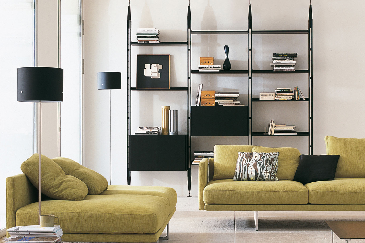 Modernes Wohnzimmer mit Sofa und passender Liege, davor eine schwarze Stehleuchte. Im Hintergrund steht ein hohes schwarzes Regal mit Büchern und Dekorobjekten an einer hellen Wand.