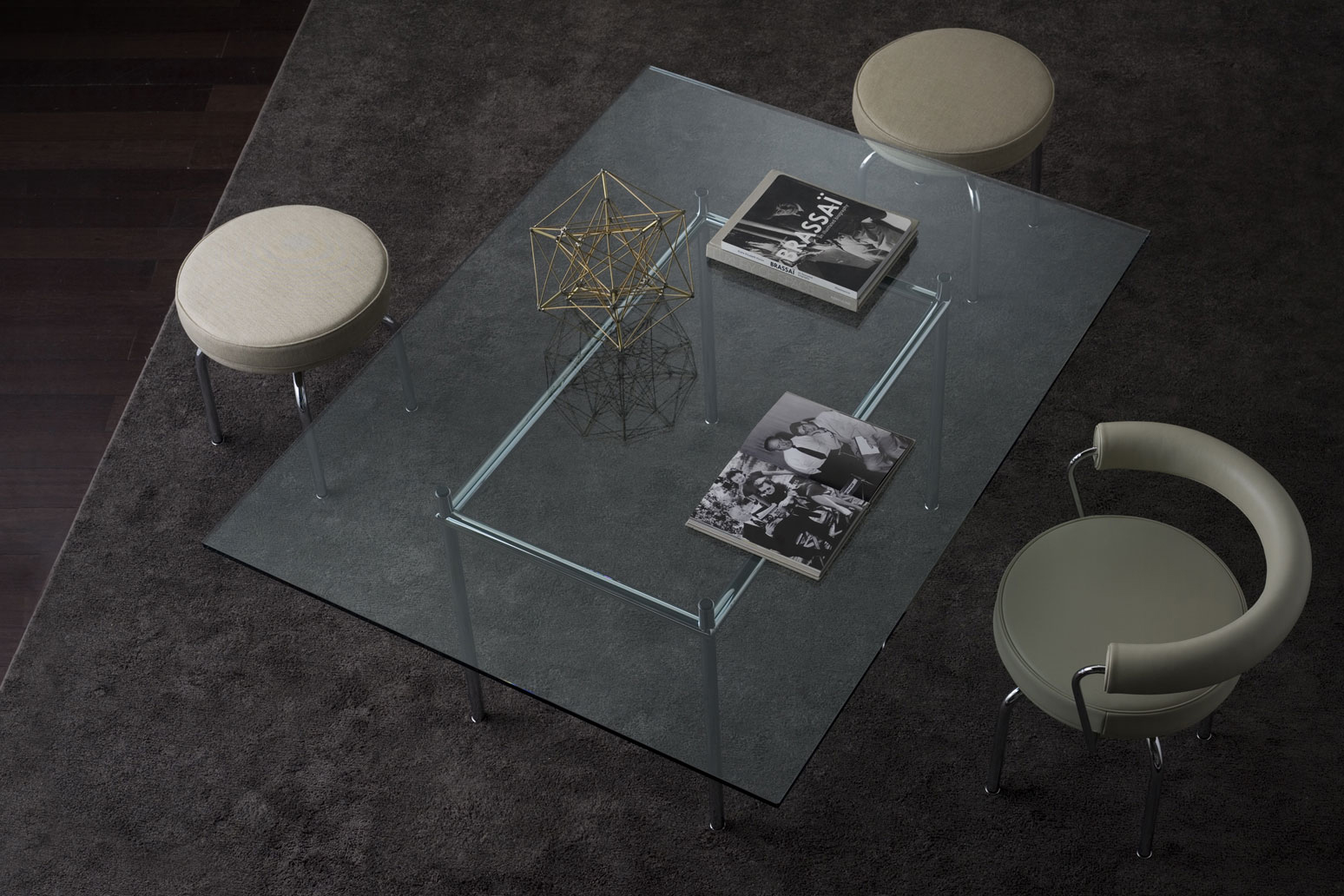Gläserner Couchtisch auf dunkelgrauem Teppich, von oben fotografiert, mit Metallrahmen, zwei Bildbänden und einer goldenen Drahtskulptur. Rundherum stehen zwei runde Hocker und ein gepolsterter Stuhl mit Armlehnen.