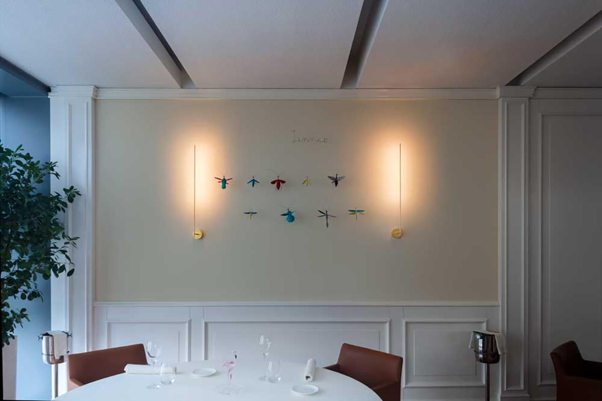 Helle Esszimmer Szene mit weisser Wandvertäfelung und zwei schlanken Wandlampen, dazwischen eine Reihe bunter Libellen Dekorationen, im Vordergrund ein weiss gedeckter Tisch mit braunen Stühlen und links eine grosse Zimmerpflanze.