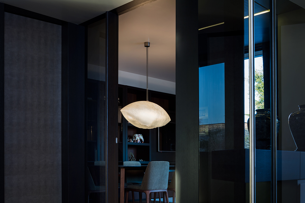 Dunkler Innenraum mit glänzenden Wandpaneelen und blauen Fensterreflexionen, zentral eine organisch geformte Pendelleuchte in warmem Licht über einem Tisch mit Stuhl und Regalnische im Hintergrund.