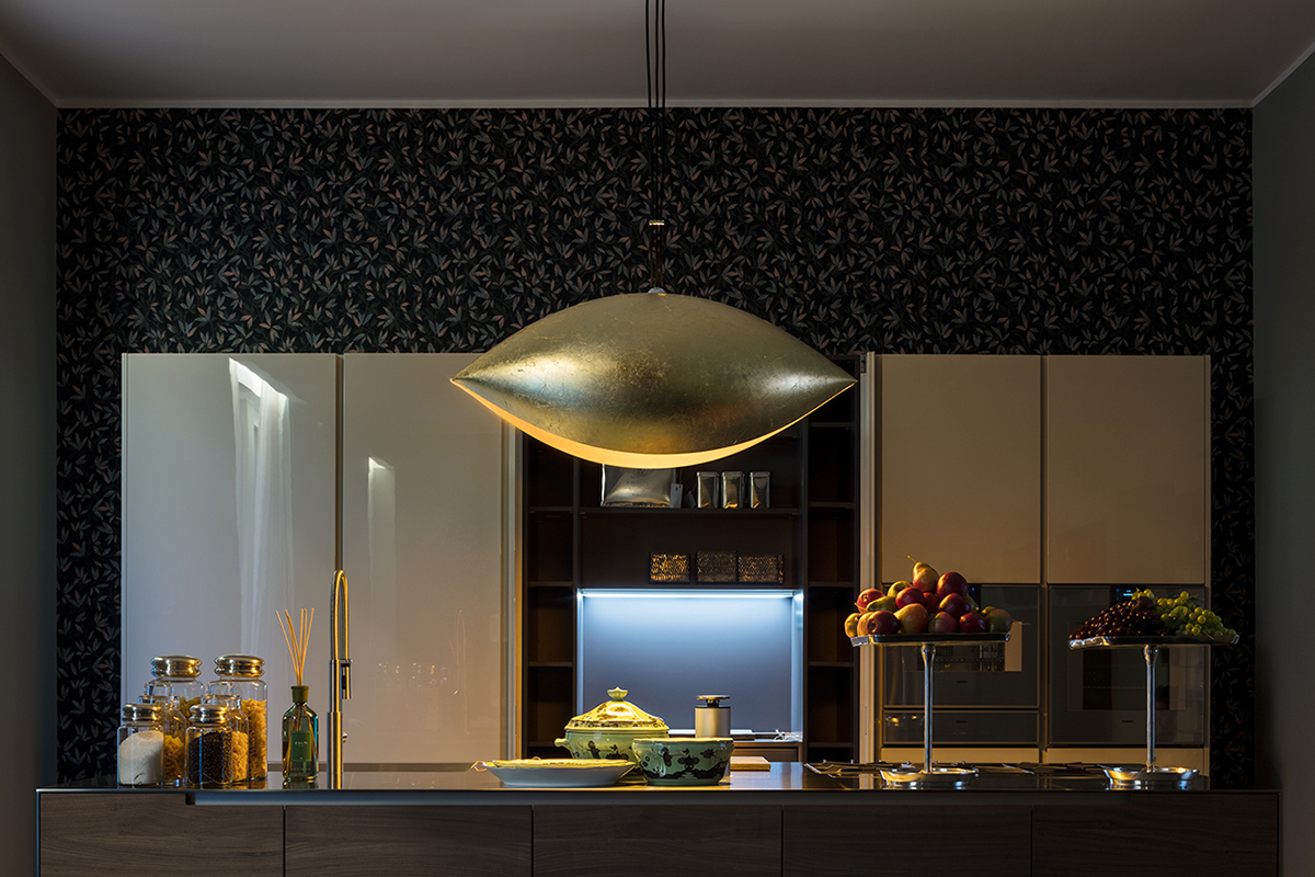 Moderne Küche mit dunkler gemusterter Tapete, glatten Schrankfronten und zentraler goldener Pendelleuchte über der Kücheninsel. Auf der Arbeitsplatte stehen Glasbehälter, Geschirr und Obstschalen, im Hintergrund ist eine beleuchtete Nische zu sehen.