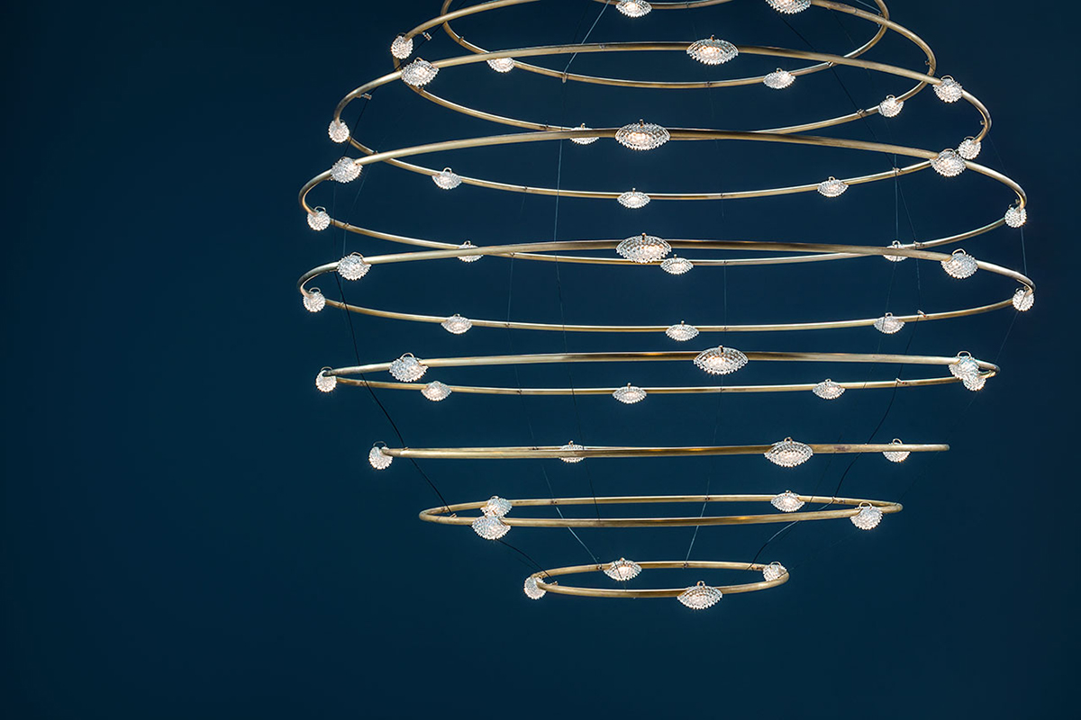 Skulpturale Hängeleuchte mit mehreren goldfarbenen, spiralförmig angeordneten Ringen und kleinen, kristallartigen Lichtpunkten vor dunkelblauem Hintergrund.