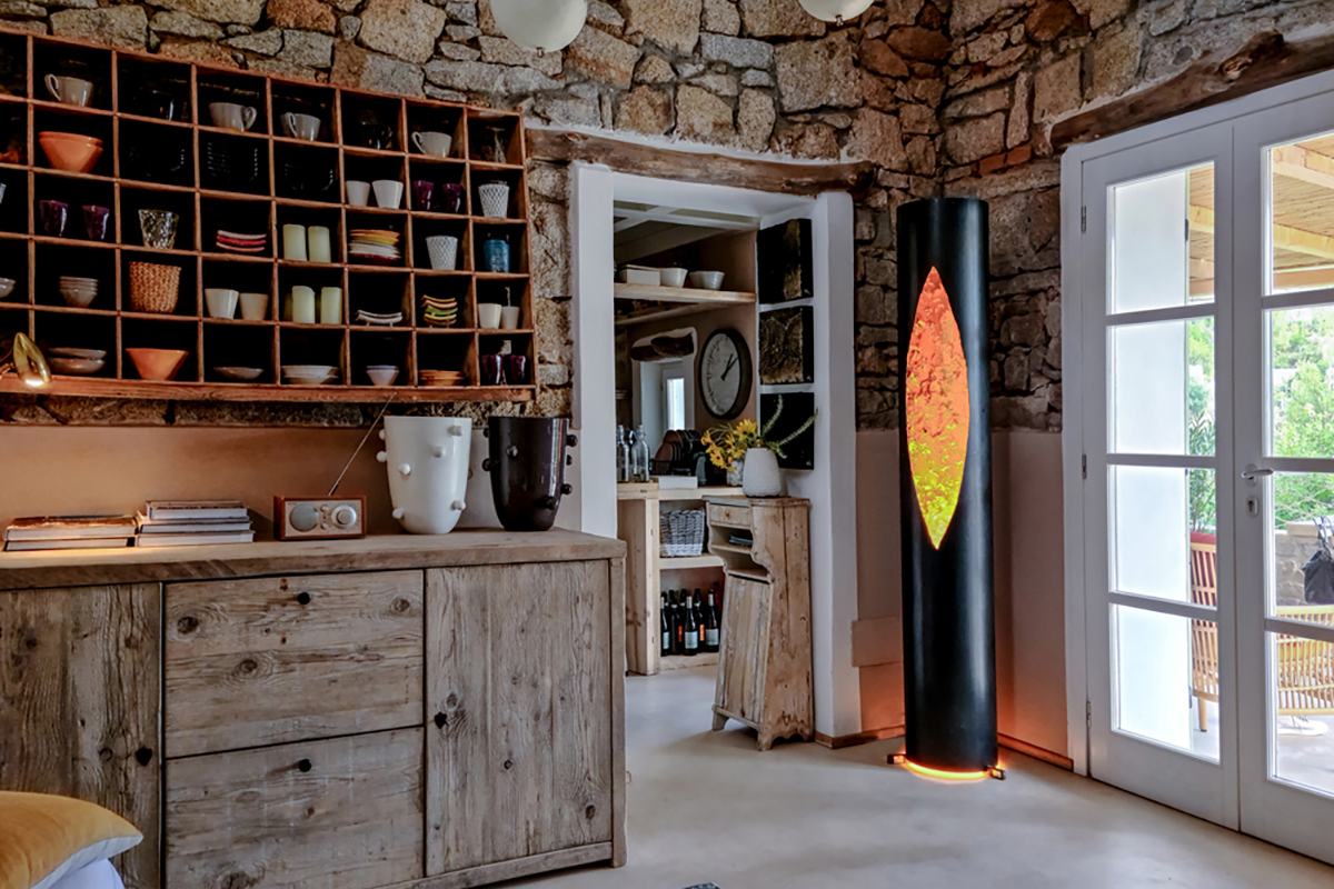 Rustikaler Raum mit Natursteinwänden und Holzmöbeln, offenes Regal voller Geschirr und Schalen, daneben hohe schwarze Stehleuchte mit leuchtendem, orangefarbenem Einsatz bei einer verglasten Türe.