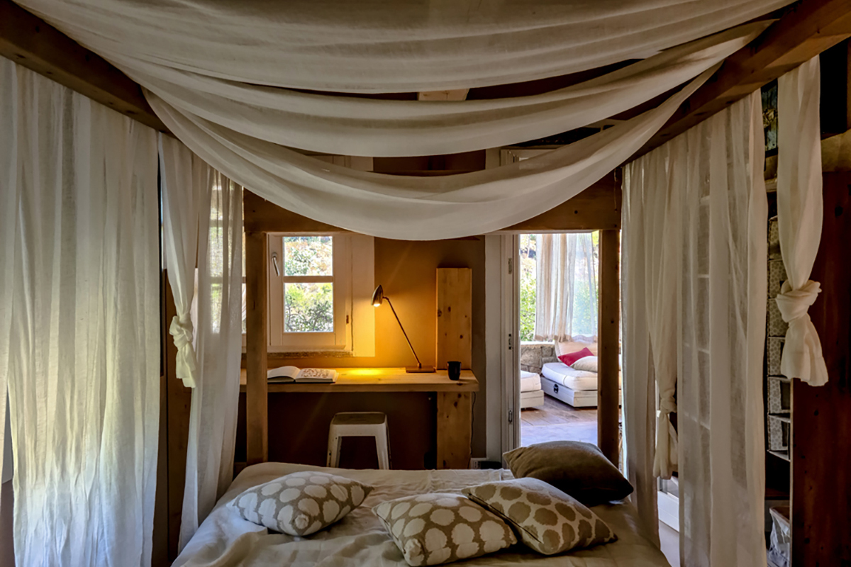 Gemütlicher Schlafbereich mit Himmelbett und drapierten Stoffbahnen, Holzverkleidung, Vorhangen und kleinem Schreibtisch mit Lampe am Fenster.