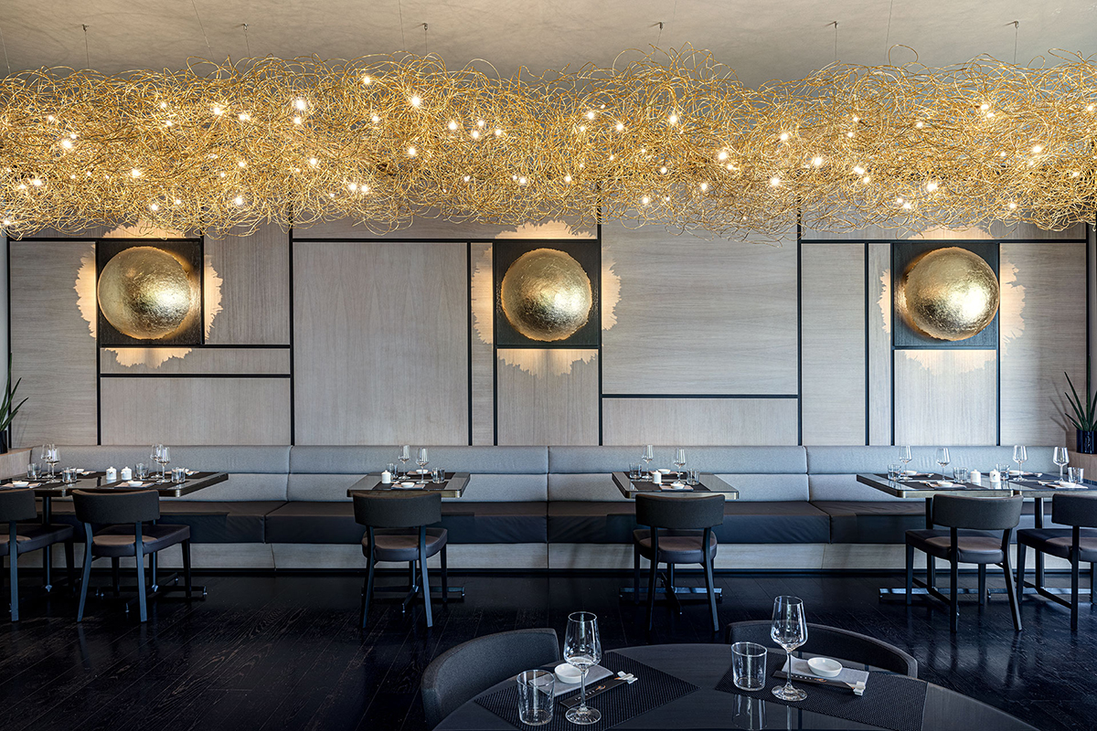 Modernes Restaurantinterieur mit langer grauer Polsterbank, gedeckten Tischen und einer grossen goldenen Lichtinstallation unter der Decke.