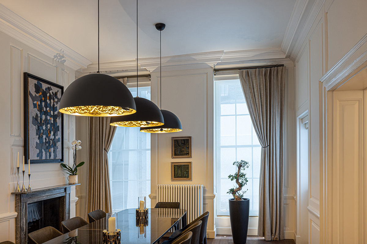 Drei schwarze Kuppel Pendelleuchten mit goldener Innenseite hängen über einem langen dunklen Esstisch in einem klassischen Raum mit Kamin, hohen Fenstern und Vorhängen.