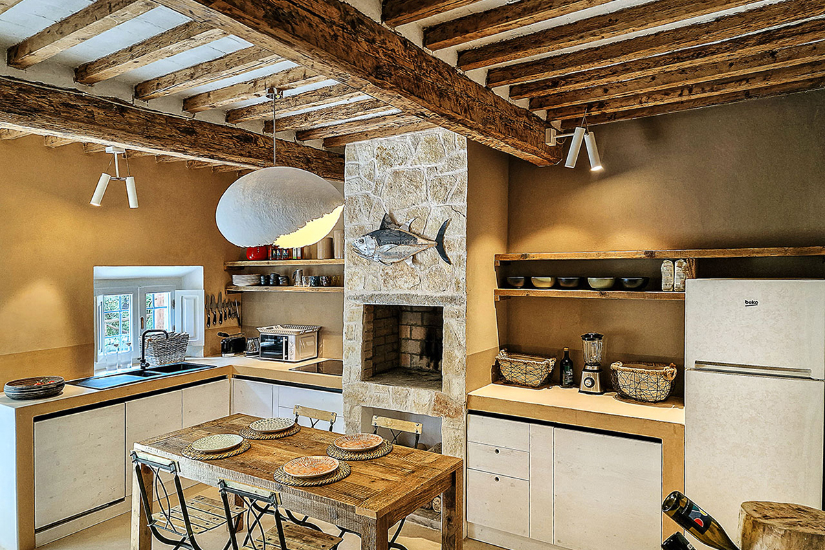 Rustikale Landhausküche mit sichtbaren Holzbalken und gemauertem Steinkamin, davor ein Holztisch mit gedeckten Tellern, weisse Einbauzeile, offene Regale und Kühlschrank.