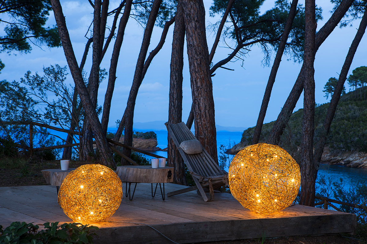 Terrasse im Pinienwald mit Blick aufs Meer bei Dämmerung, zwei leuchtende Kugelleuchten aus Geflecht, dazu Holzliege und niedrige Tische.