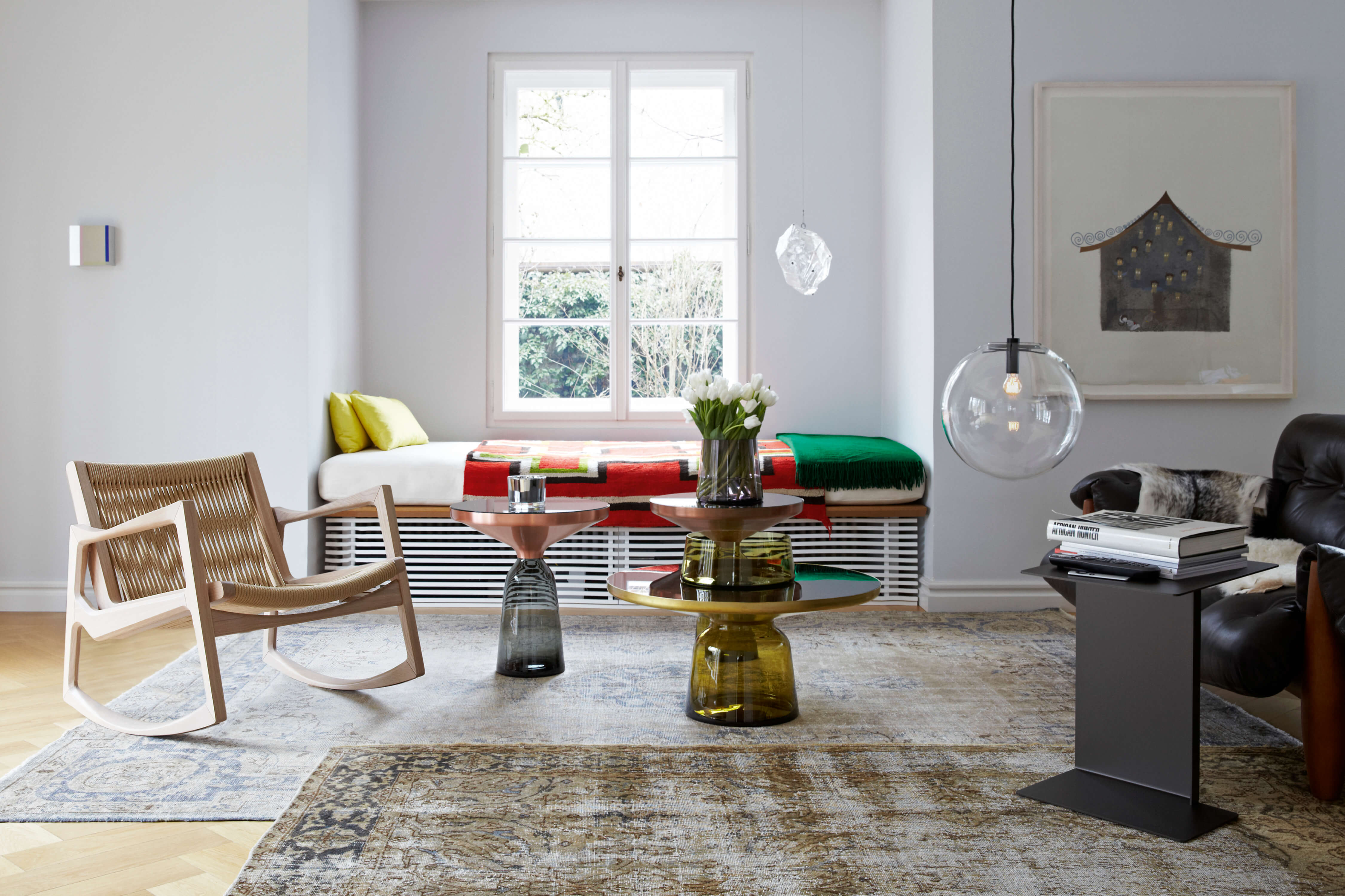 Heller Wohnraum mit Fensterbank Sitzplatz. Links ein Holz Schaukelstuhl mit geflochtener Sitzfläche. Unter dem Fenster eine Polsterbank mit buntem Plaid und grüner Decke. In der Mitte zwei runde Beistelltische aus Glas mit Vase weisser Tulpen. Rechts eine grosse Glas Pendelleuchte und ein Beistelltisch mit Büchern auf gemustertem Teppich.
