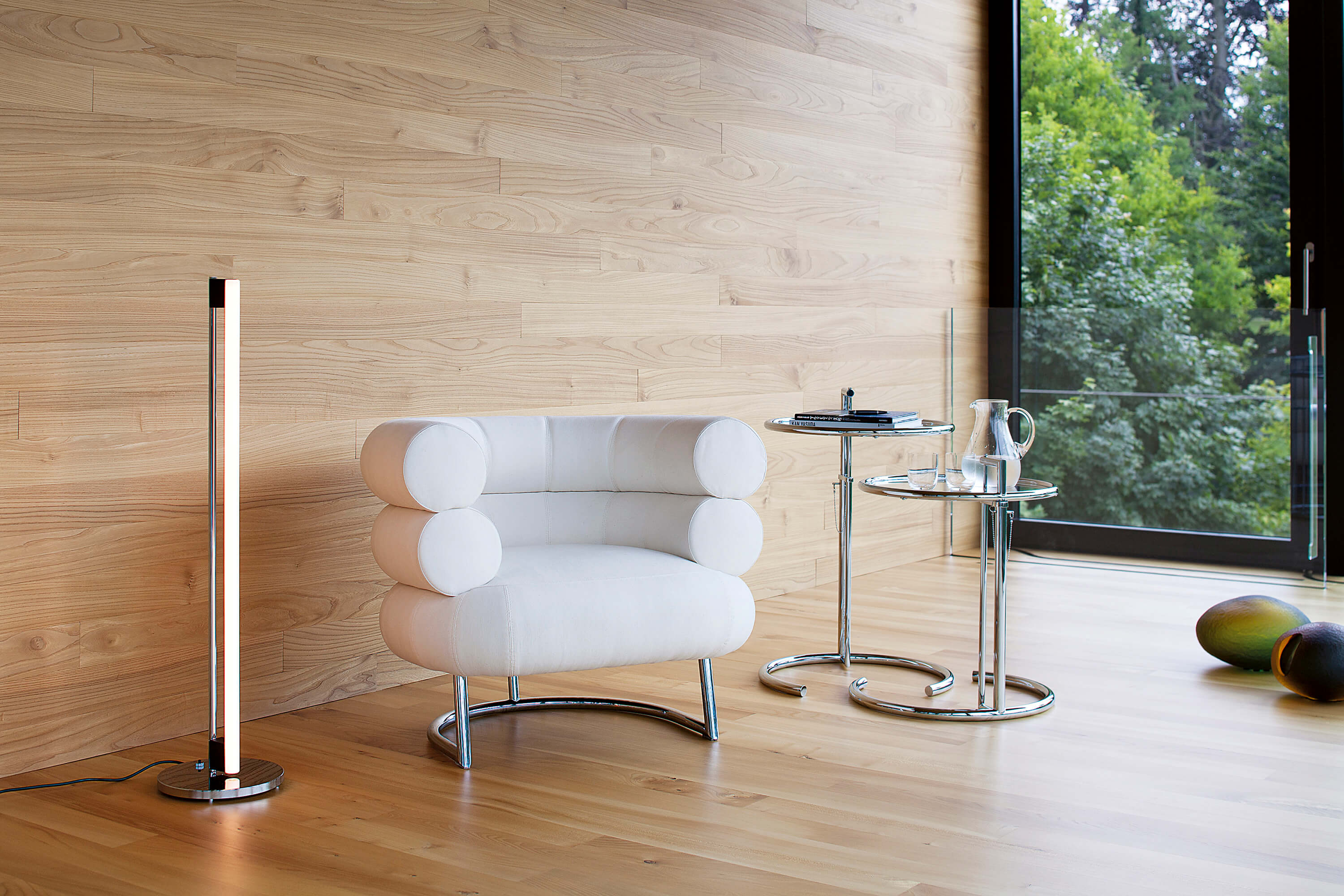 Minimalistische Lounge Ecke mit hellem Holz. Weisser Polstersessel mit zylindrischen Armlehnen und Chromgestell. Daneben zwei runde Beistelltische aus Chrom und Glas mit Büchern, Karaffe und Gläsern. Links eine schlanke Stehleuchte, rechts ein grosses Fenster mit Blick ins Grüne.