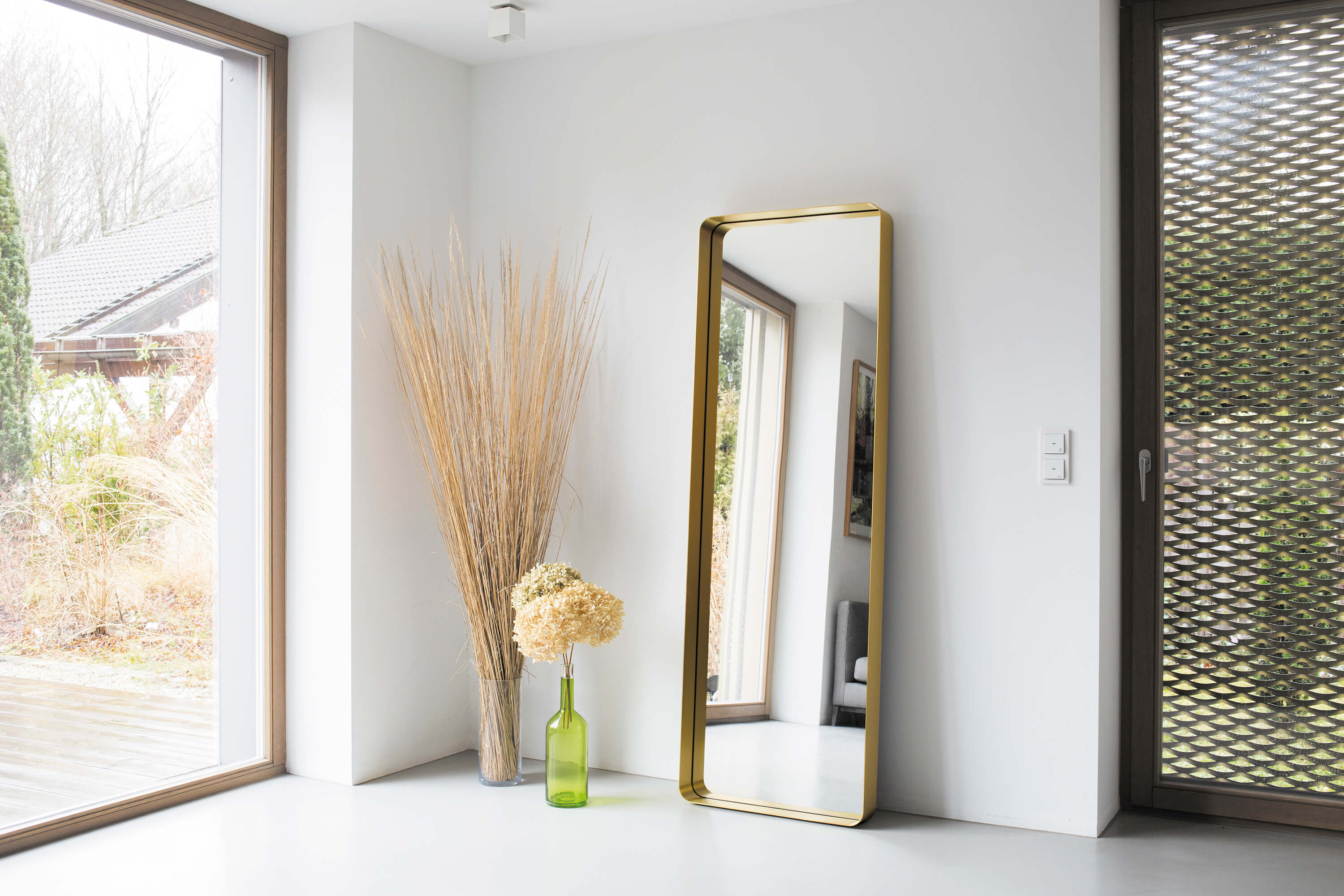 Heller Eingangsbereich mit bodentiefen Fenstern und einem grossen Standspiegel mit goldfarbenem Rahmen, der an der Wand lehnt. Daneben stehen eine hohe Vase mit Trockengras sowie eine grüne Glasflasche mit heller Hortensie.