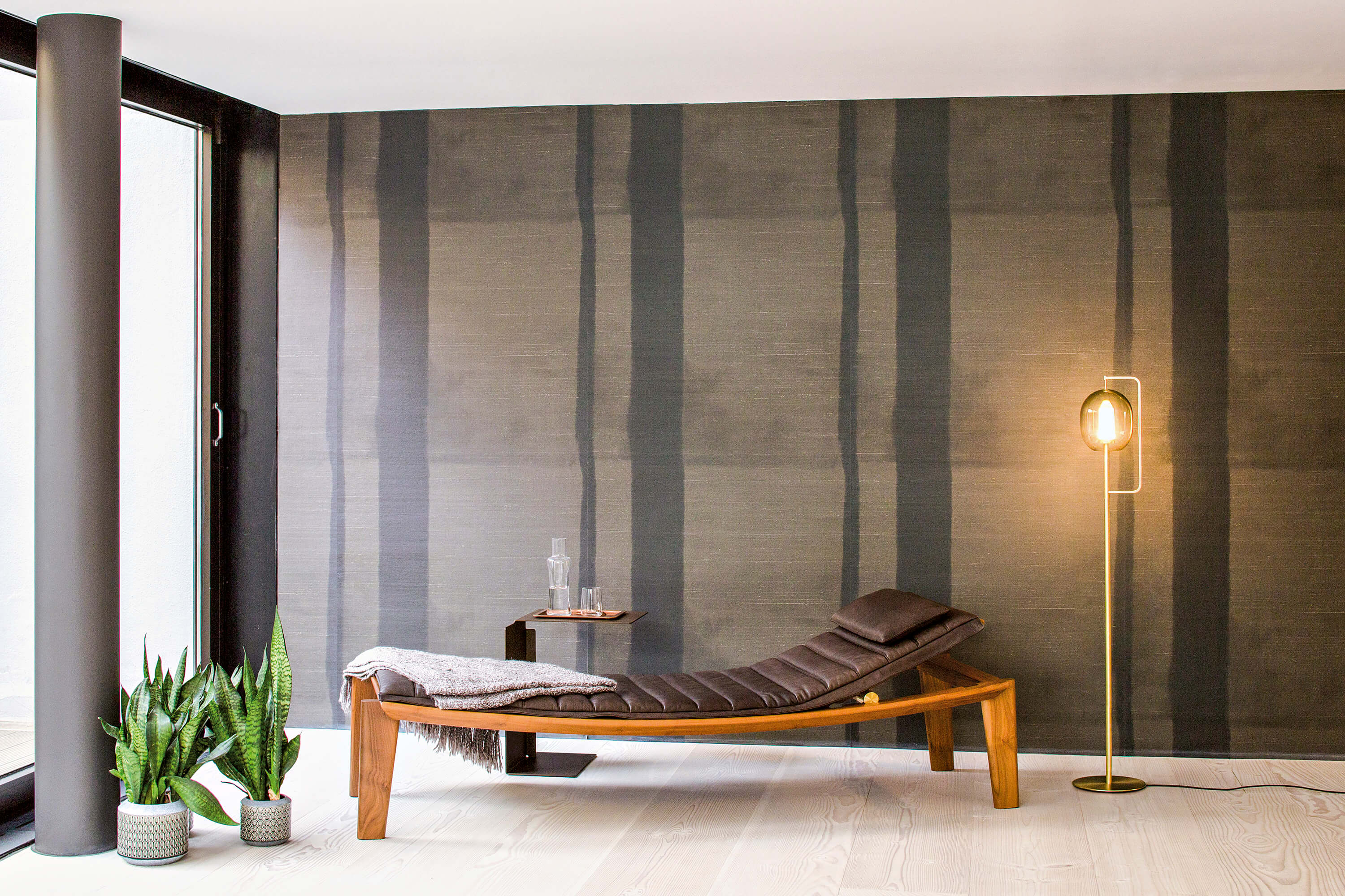 Minimalistischer Ruhebereich mit Holz Liege und dunkelbraunem Polster vor grauer Wand mit vertikalen Streifen. Links grosse Fensterfront und zwei Topfpflanzen. Neben der Liege ein kleiner Beistelltisch mit Karaffe und Gläsern, auf der Liege eine Decke. Rechts eine schlanke Stehleuchte mit warmem Licht.