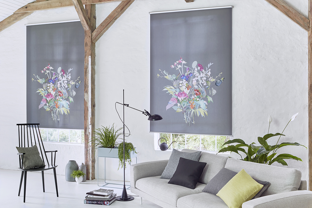 Heller Wohnraum mit Sichtbalken, grauem Sofa und schwarzem Stuhl. Zwei graue Rollos mit farbigem Blumenmotiv vor den Fenstern, dazu Stehleuchte und mehrere Zimmerpflanzen.