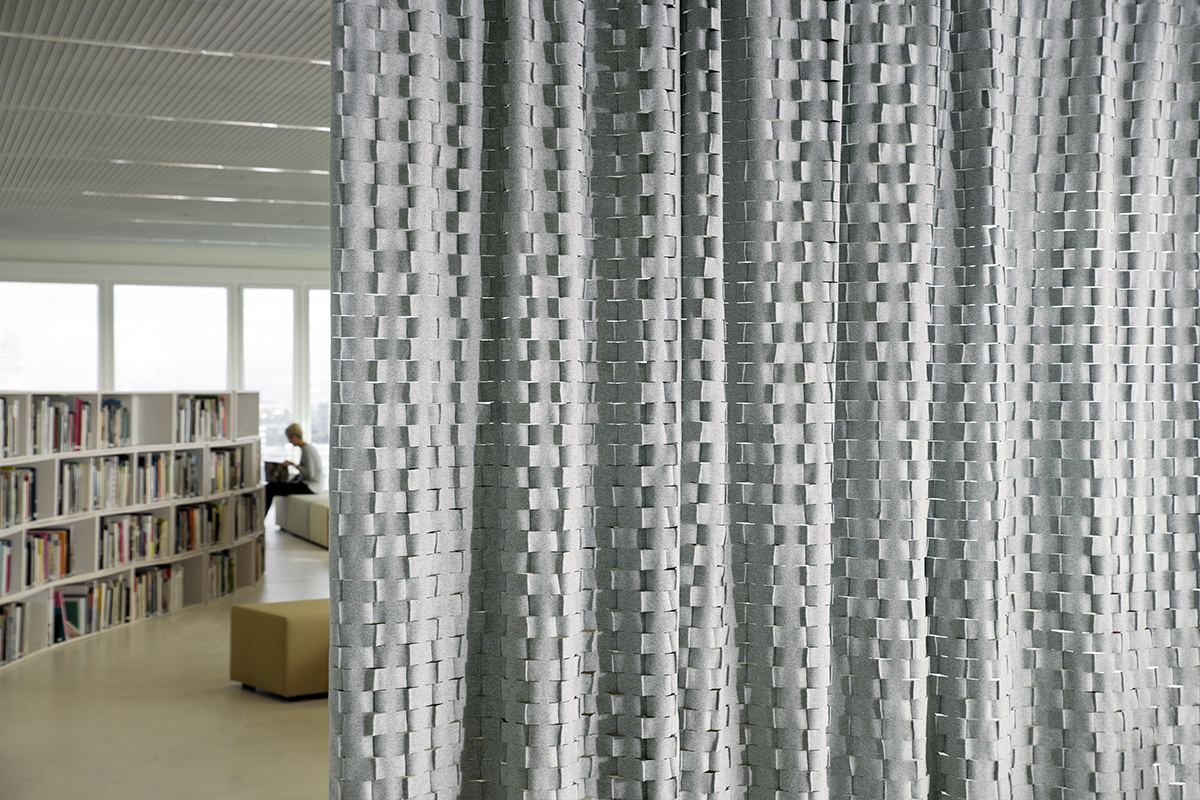 Grauer, strukturierter Vorhang aus vielen kleinen, geflochten wirkenden Elementen im Vordergrund. Im Hintergrund eine helle Bibliothek mit Bücherregalen, grossen Fenstern und einer Person, die auf einer Bank liest.