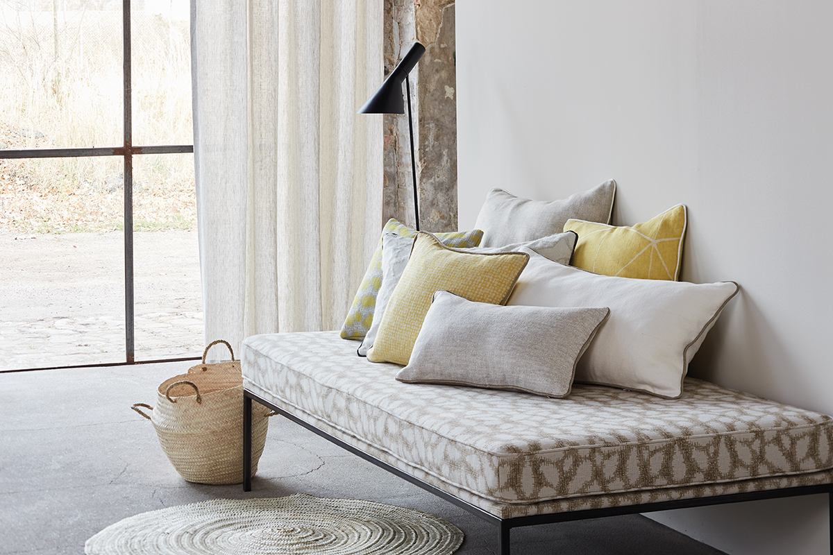 Helle Daybed mit gemustertem Bezug und mehreren Kissen in Grau, Weiss und Gelb. Daneben ein schwarzer Stehleuchter und ein leichter Vorhang vor grossem Fenster. Auf dem Boden stehen geflochtene Körbe und ein runder Teppich auf Betonboden.