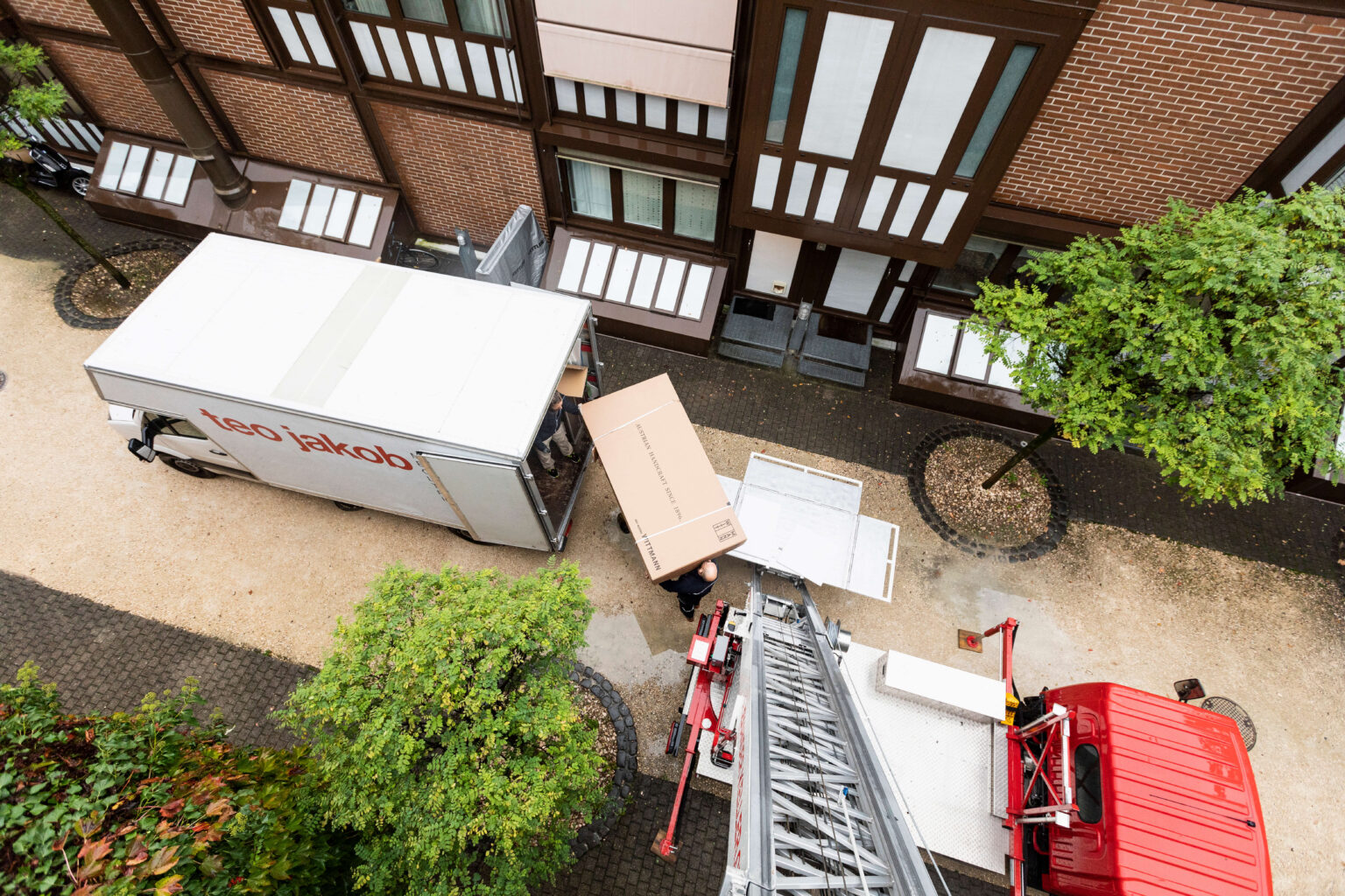 Weisser Lieferwagen mit rotem teo jakob Logo und dem Schriftzug Einrichtung und Innenarchitektur, teilweise von grünem Efeu im Vordergrund verdeckt, vor einem Gebäude.
