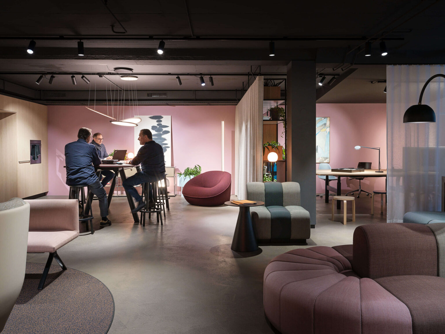Showroom Szene mit drei Personen an einem Hochtisch, umliegenden Sitzmöbeln und pendelnden Leuchten vor einer rosafarbenen Wand.