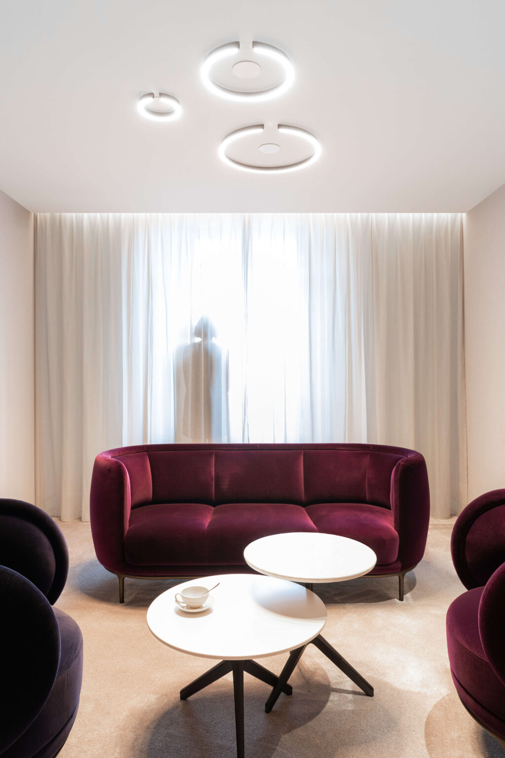 Kundenlounge in Genf mit bordeauxfarbenen Samtsofas, runden Deckenleuchten und Tageslichtvorhang.