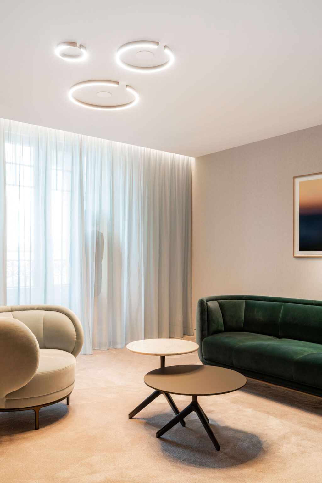 Kundenlounge mit samtbezogenen Sesseln und ringförmigen Occhio-Deckenleuchten vor leichten Vorhängen in einer Privatbank in Genf.