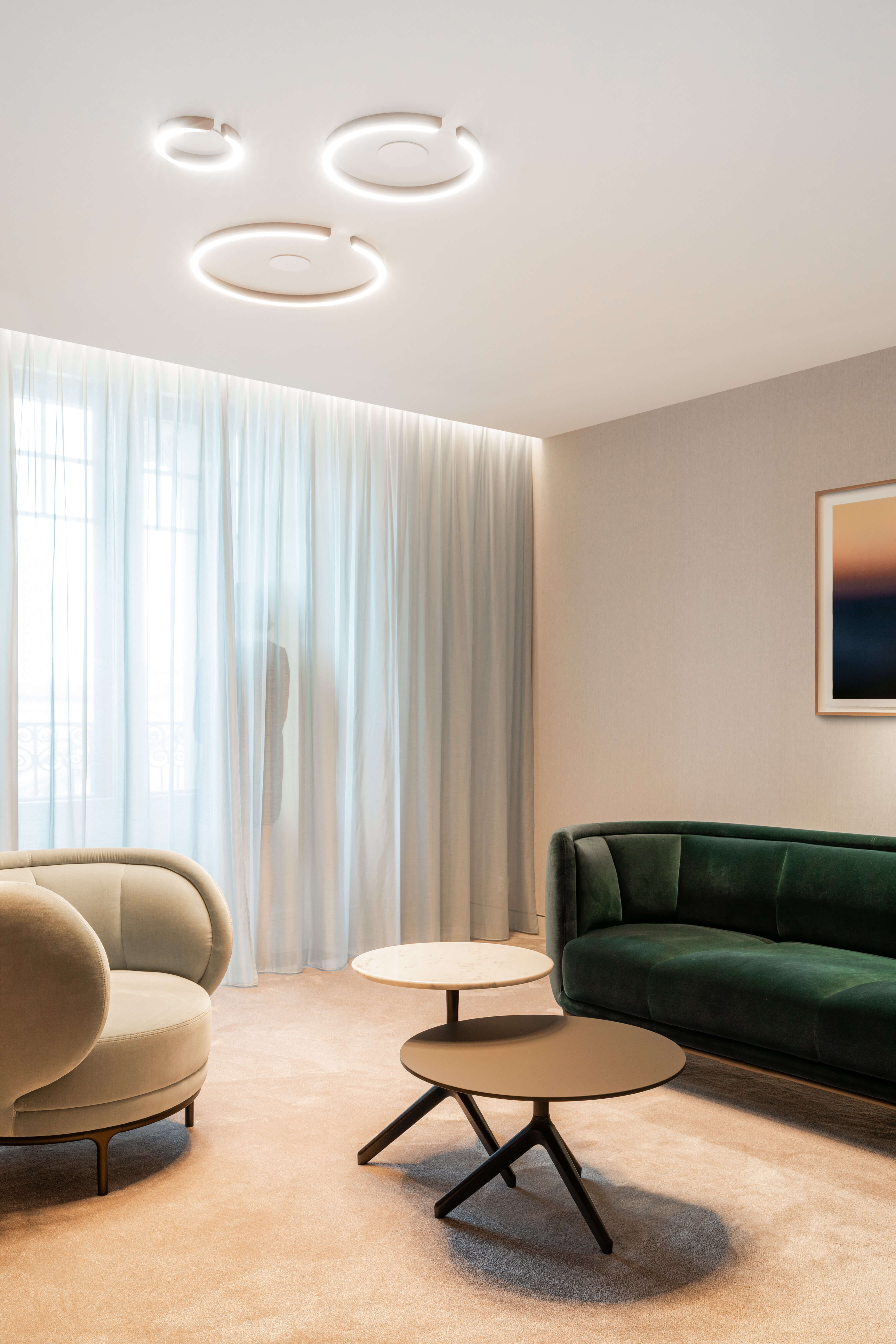 Kundenlounge mit samtbezogenen Sesseln und ringförmigen Occhio-Deckenleuchten vor leichten Vorhängen in einer Privatbank in Genf.