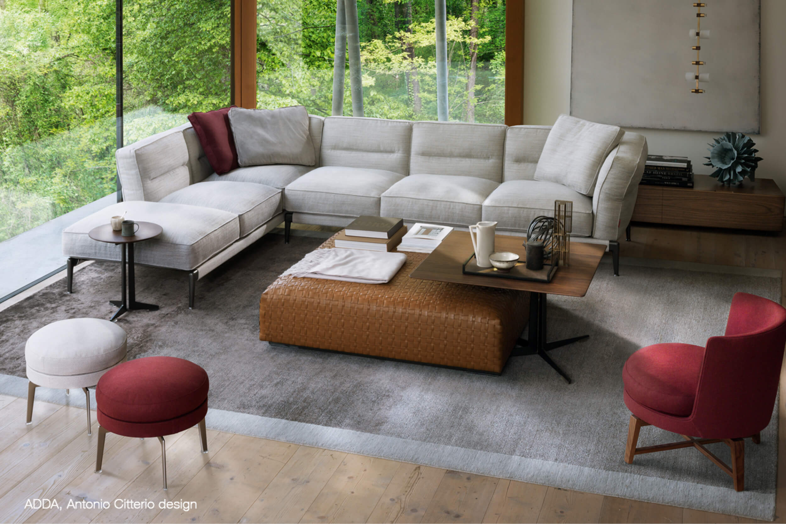 Helles Wohnzimmer mit grauem Ecksofa, grossem Lederhocker und Beistelltisch auf Teppich vor bodentiefem Fenster mit Grünblick.