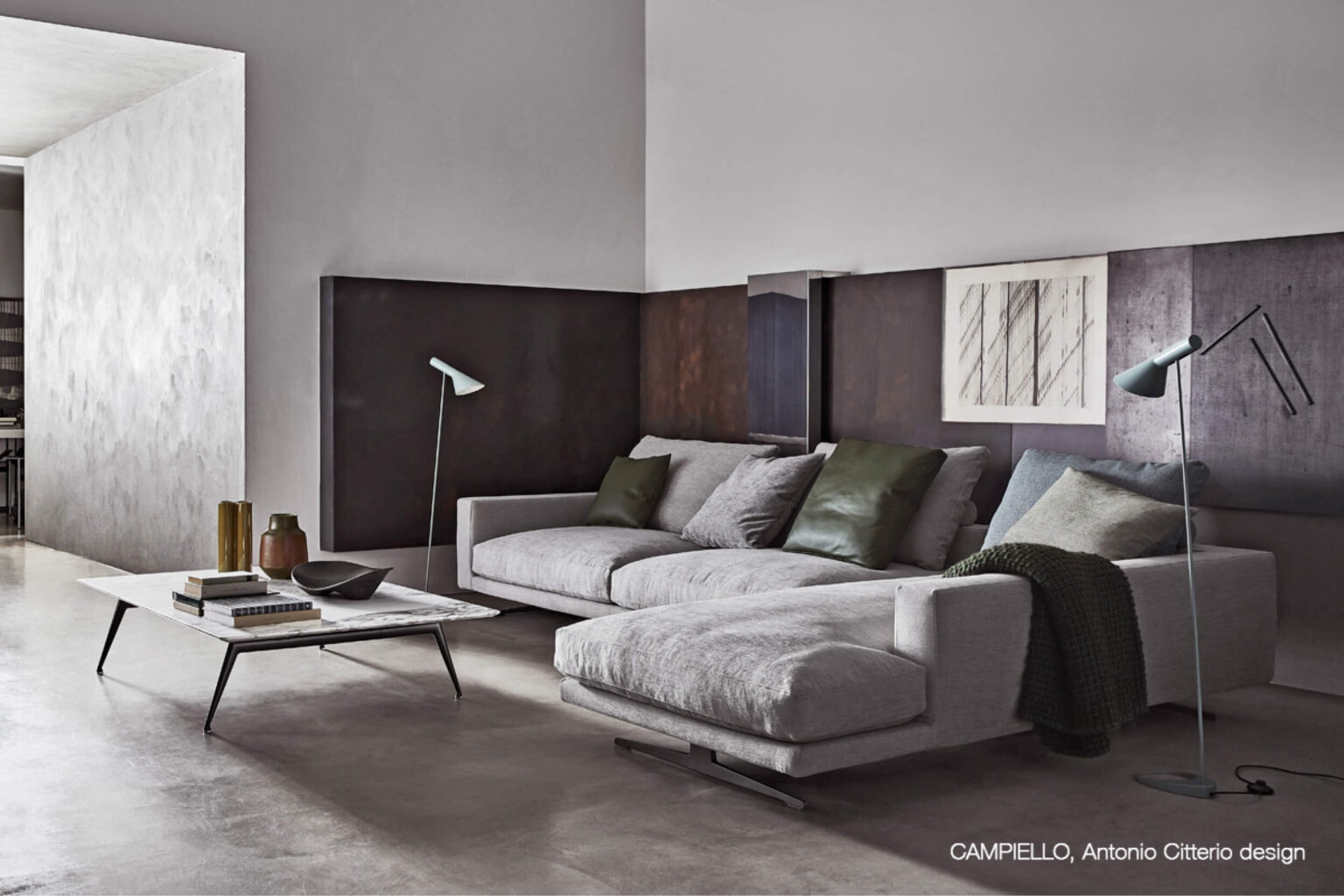 Modernes Wohnzimmer mit grossem hellgrauem Ecksofa und Chaiselongue vor dunklen Wandpaneelen. Auf dem Sofa liegen grüne und graue Kissen, daneben stehen zwei schlanke Stehleuchten und davor ein niedriger Couchtisch mit Büchern und Deko.