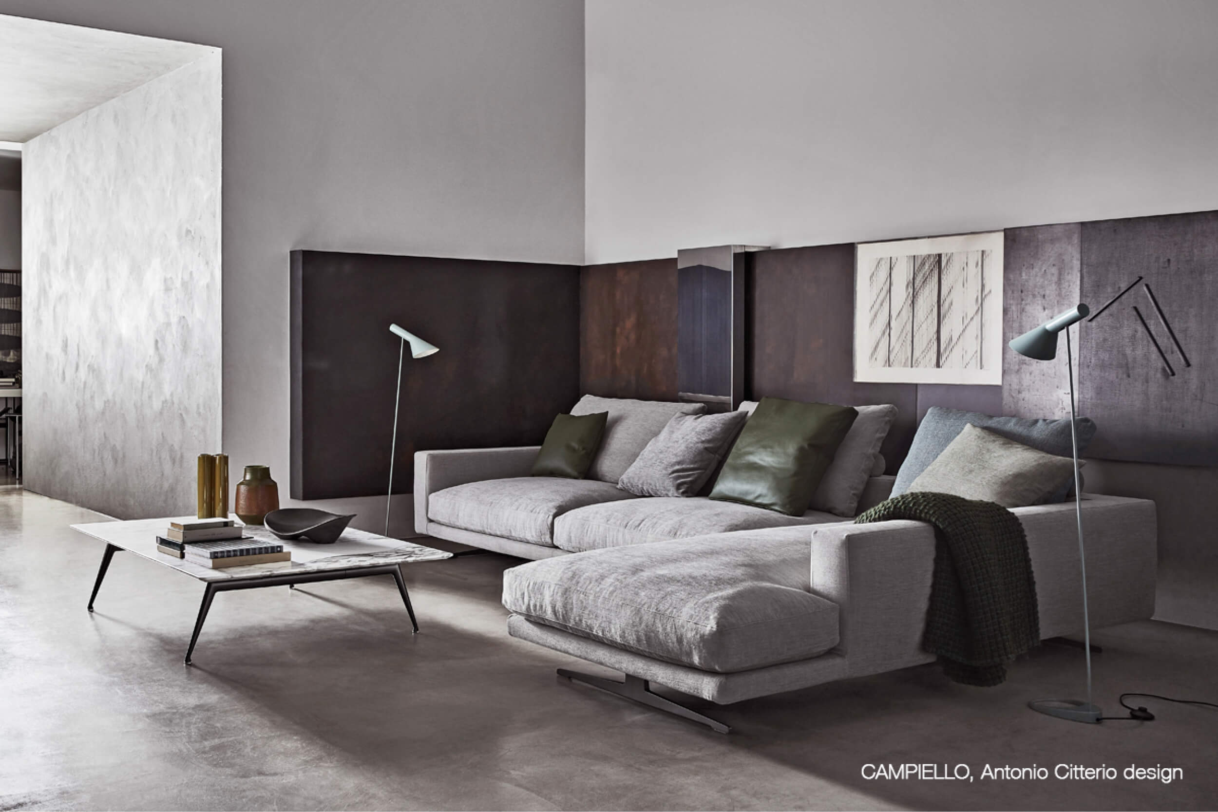 Modernes Wohnzimmer mit grossem hellgrauem Ecksofa und Chaiselongue vor dunklen Wandpaneelen. Auf dem Sofa liegen grüne und graue Kissen, daneben stehen zwei schlanke Stehleuchten und davor ein niedriger Couchtisch mit Büchern und Deko.