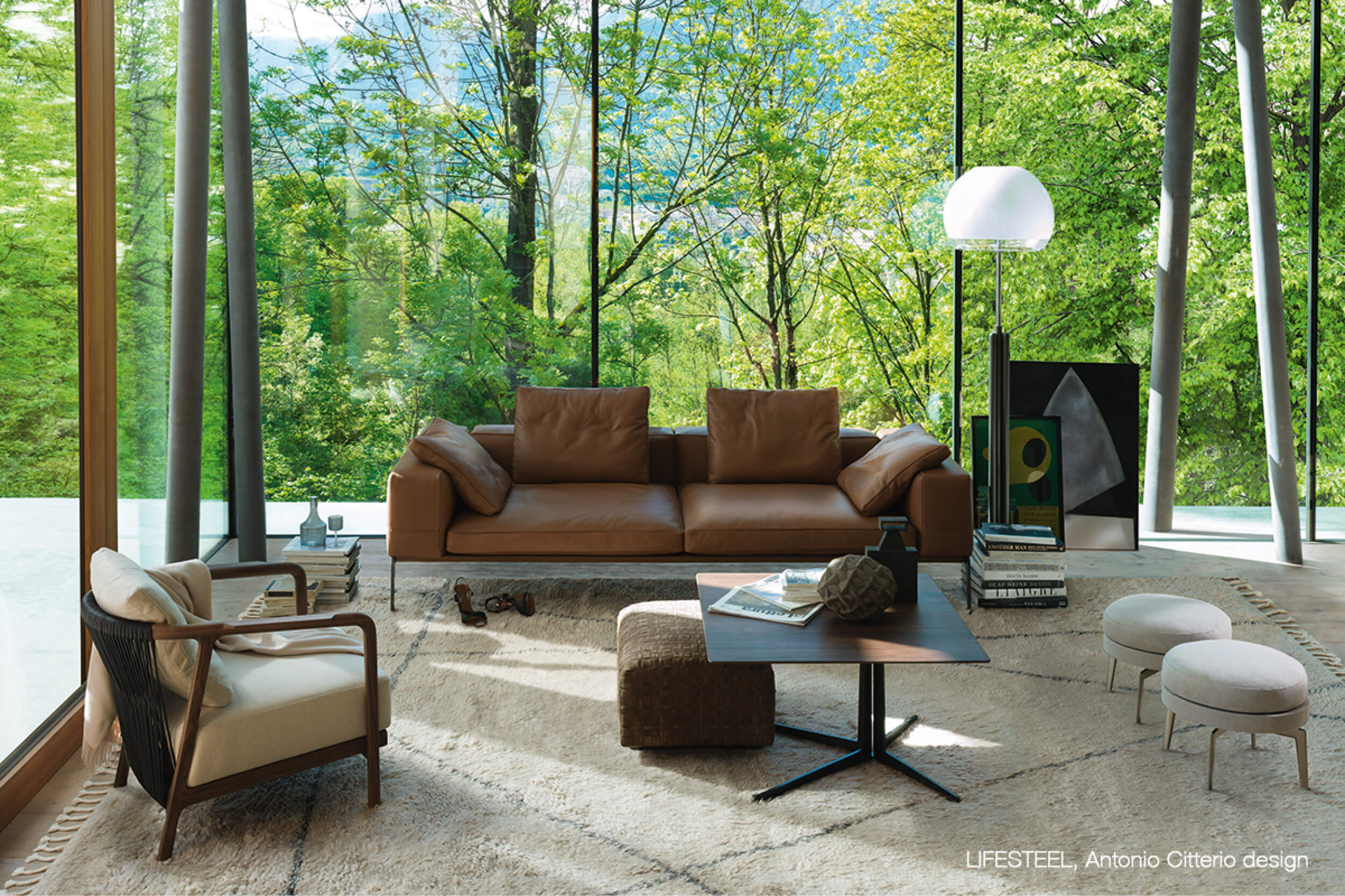 Helles Wohnzimmer mit bodentiefen Fenstern und Blick ins Grün. In der Mitte steht ein braunes Ledersofa auf einem hellen Teppich, davor ein niedriger Tisch mit Büchern und Deko. Links ein Sessel, rechts eine Stehleuchte, zwei Poufs und gerahmte Kunst an der Wand.