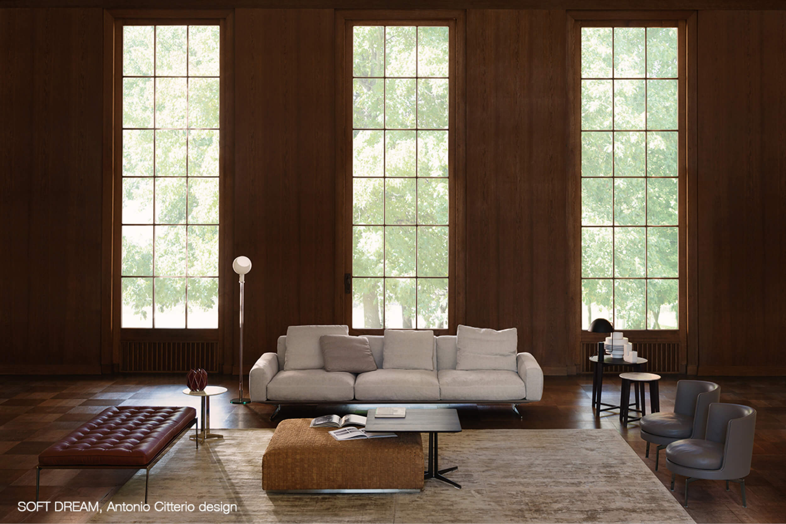 Grosses, holzvertäfeltes Wohnzimmer mit drei hohen Sprossenfenstern, hellem Sofa auf Teppich sowie Lederbank, Couchtisch und Beistelltischen mit Leuchten.