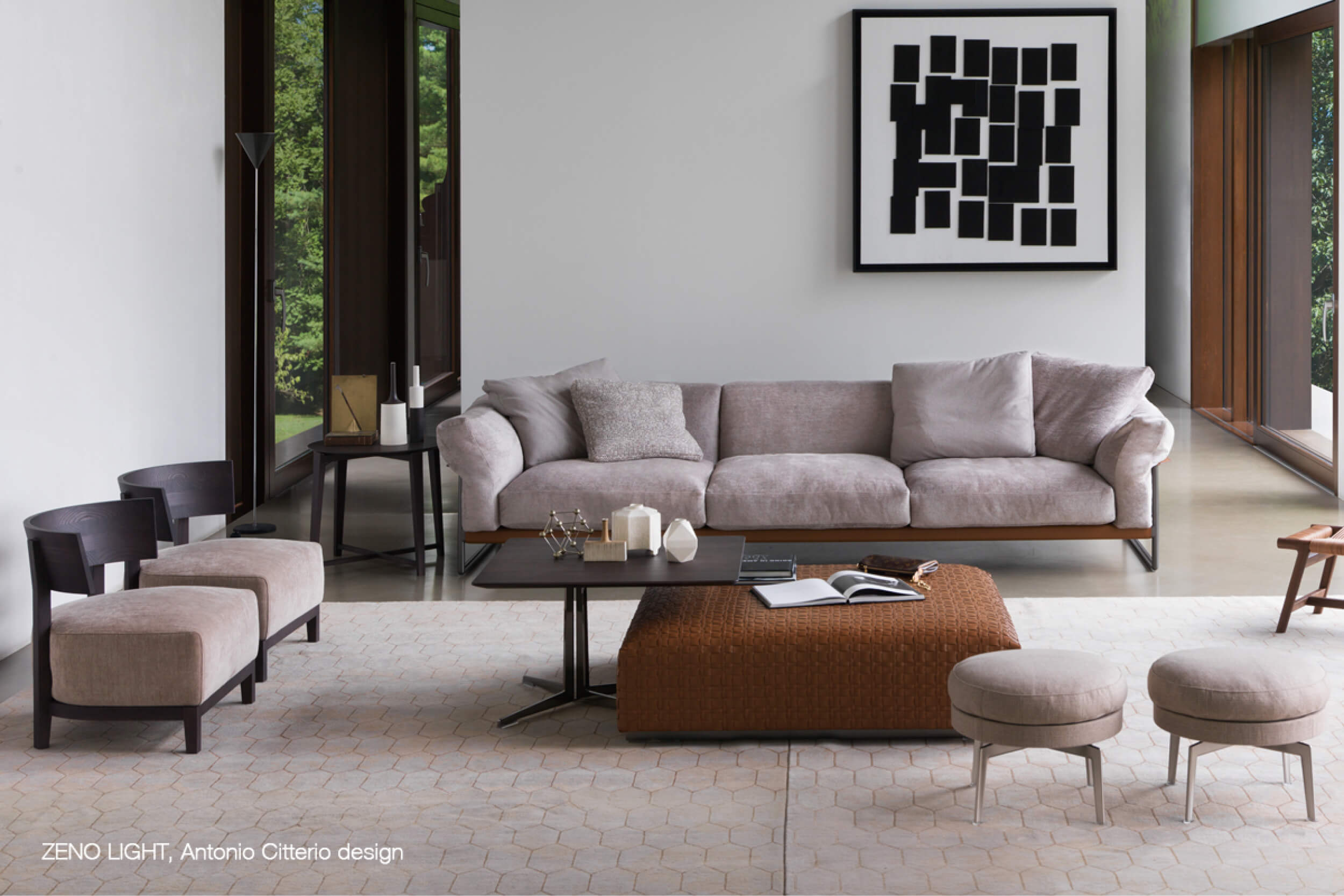 Modernes Wohnzimmer mit grauem Dreisitzer Sofa, schwarzem Couchtisch und braunem Lederhocker auf hellem Teppich, dazu Sessel und Hocker vor grossen Fenstern und grafischem Wandbild.