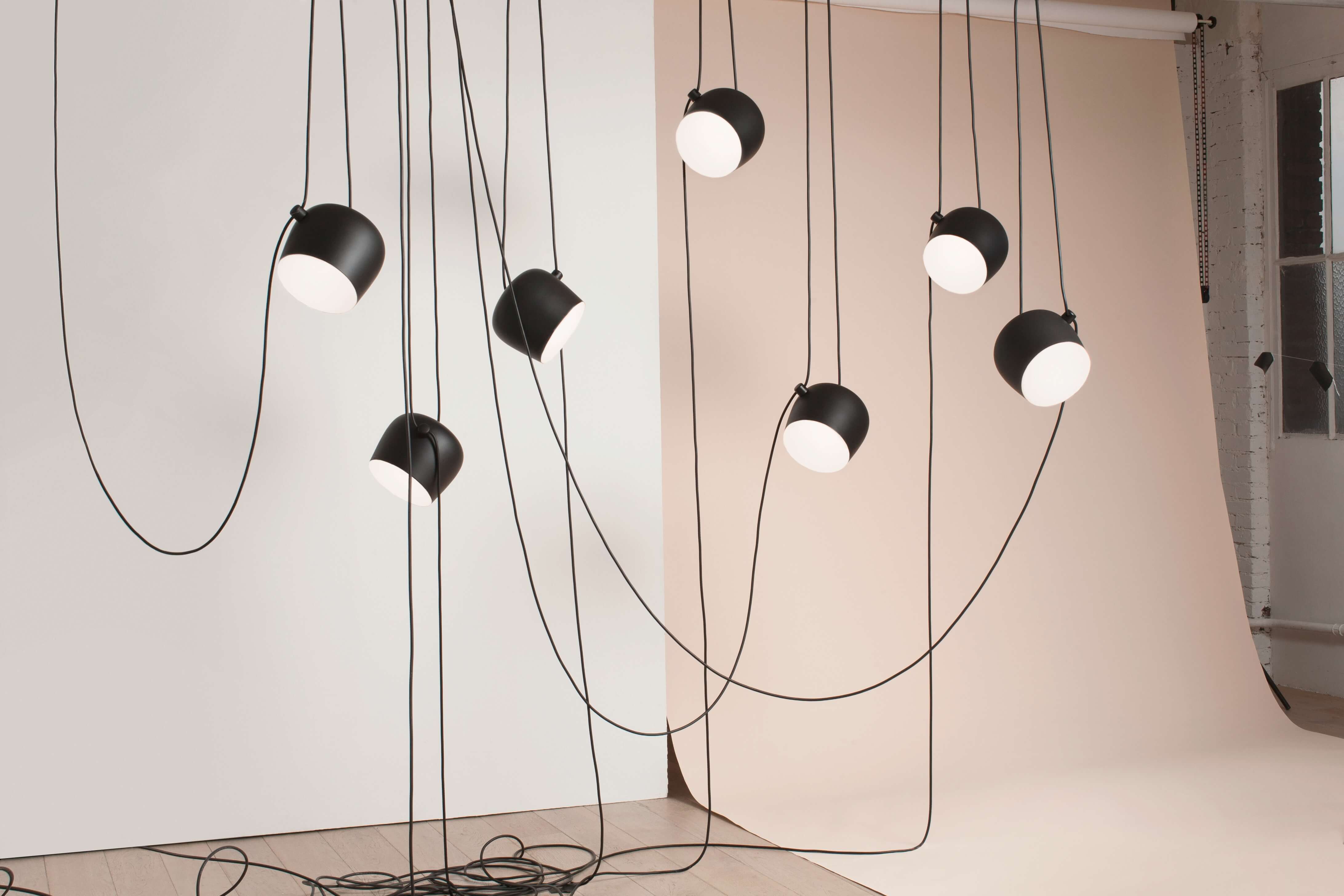 Mehrere schwarze Pendelleuchten mit runden, weissen Lichtköpfen hängen an langen Kabeln in unterschiedlichen Höhen. Die Kabel bilden eine lockere, grafische Linienführung vor hellem Hintergrund. Das Setting wirkt minimalistisch und lichtbetont.