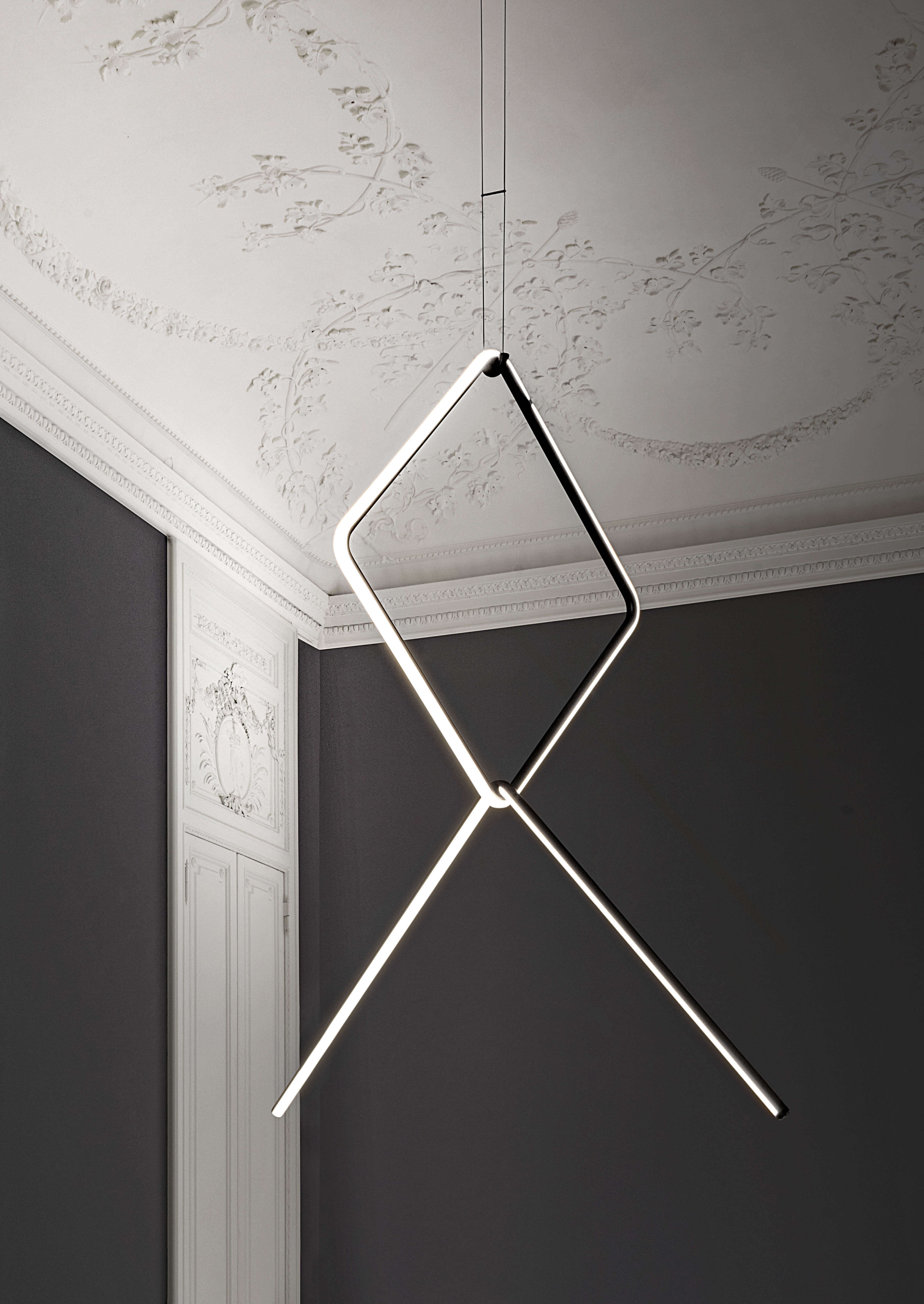 Skulpturale Pendelleuchte aus zwei leuchtenden Stäben, die eine geometrische X Form bilden. Sie hängt in einer Raumecke mit dunklen Wänden und einer reich verzierten Stuckdecke. Helles Licht setzt einen starken Kontrast zur klassischen Architektur.