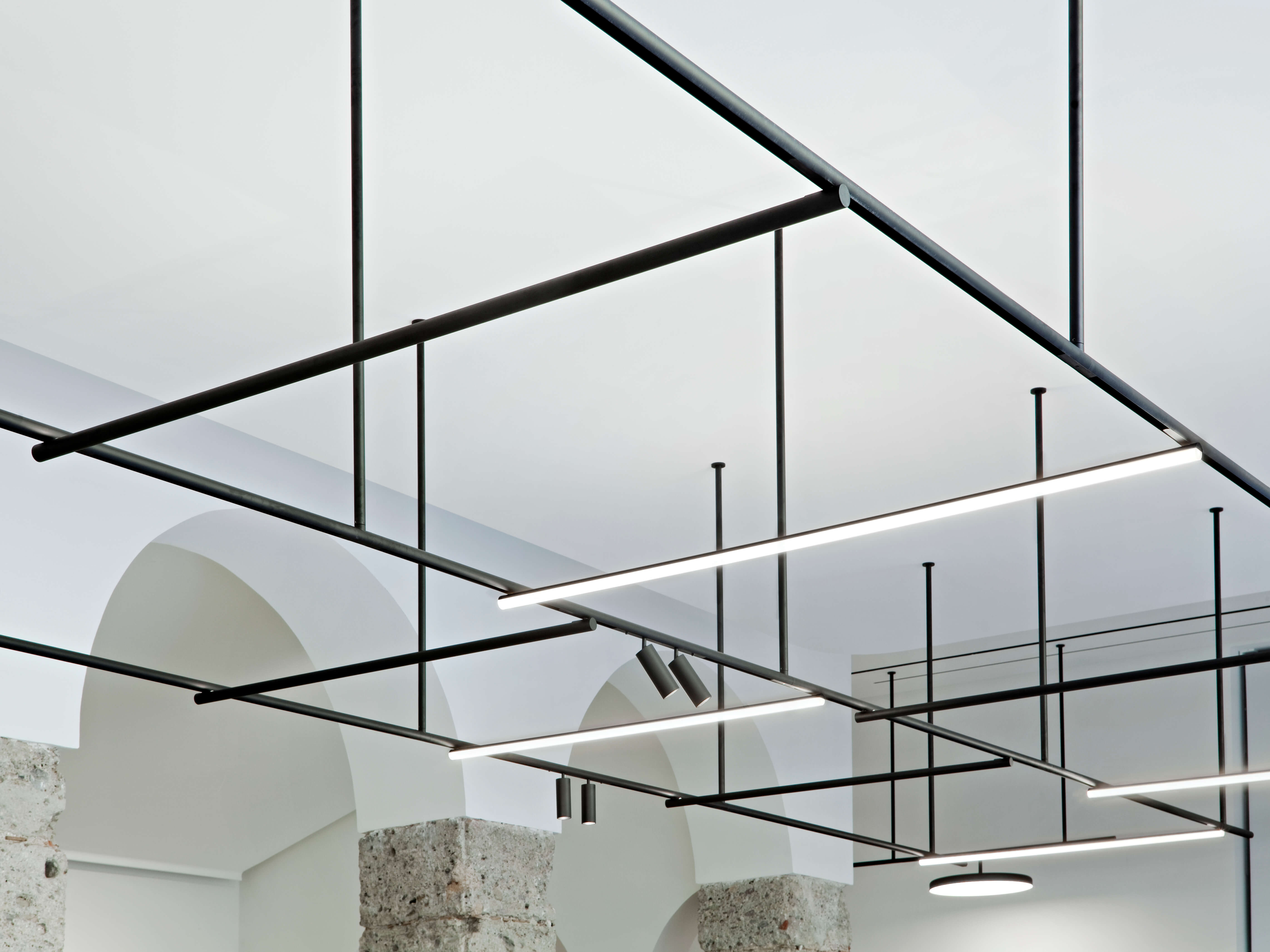 Deckenansicht mit modernem Lichtsystem aus schwarzen Rohrprofilen und linearen LED Leuchten. Mehrere Abhängungen strukturieren den Raum. Im Hintergrund sind weisse Rundbögen und Natursteinpfeiler sichtbar.