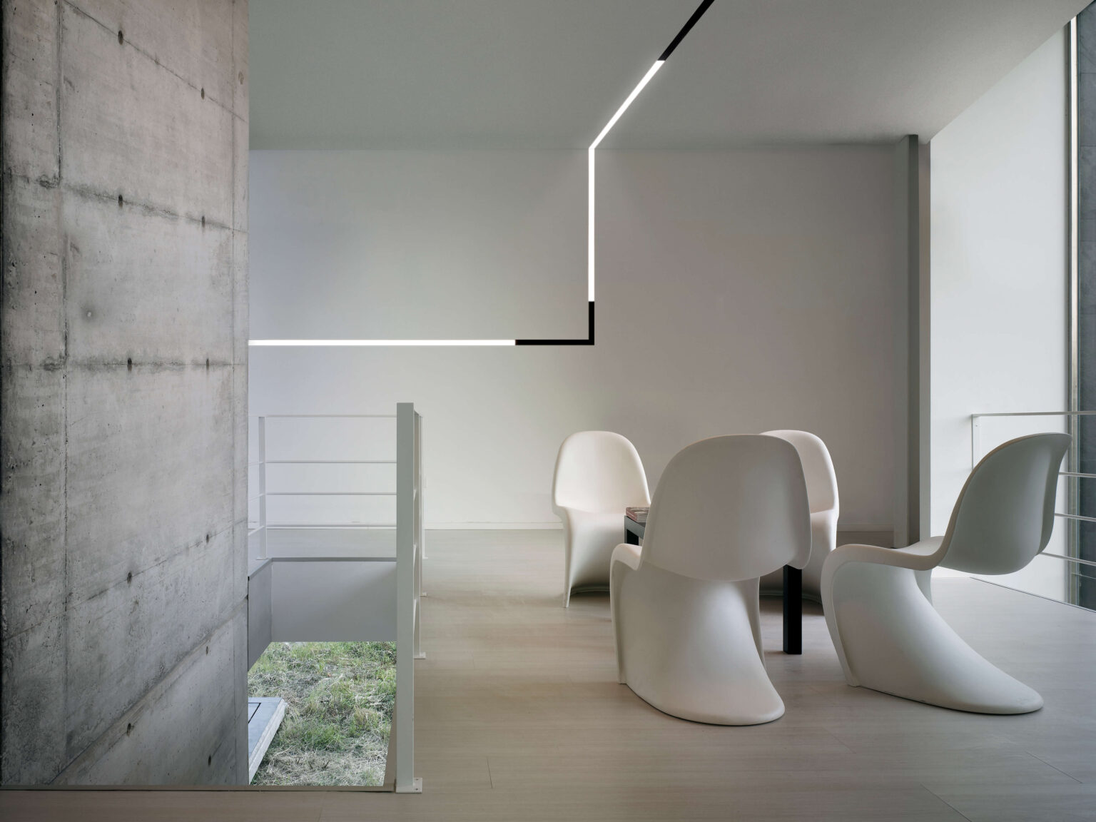 Minimalistischer Essbereich mit vier skulpturalen, weissen Stühlen um einen dunklen Tisch. Eine lineare Deckenleuchte zieht sich als L Form über die Decke. Links steht eine rohe Betonwand, rechts eine Glasfront mit Tageslicht. Heller Holzboden und schlichtes Geländer betonen die klare Architektur.